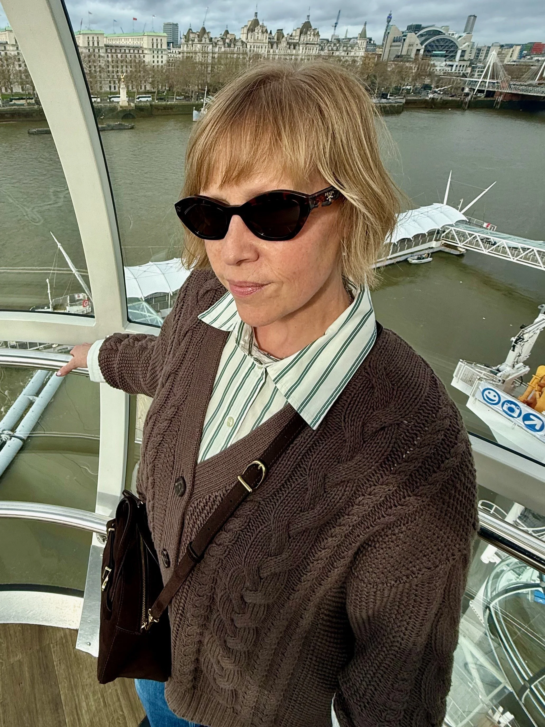 A woman with short light brown hair wearing dark sunglasses, a white shirt with green stripes, and a brown knitted cardigan, taking a selfie inside a Ferris wheel in London with the Thames River and city buildings in the background.
