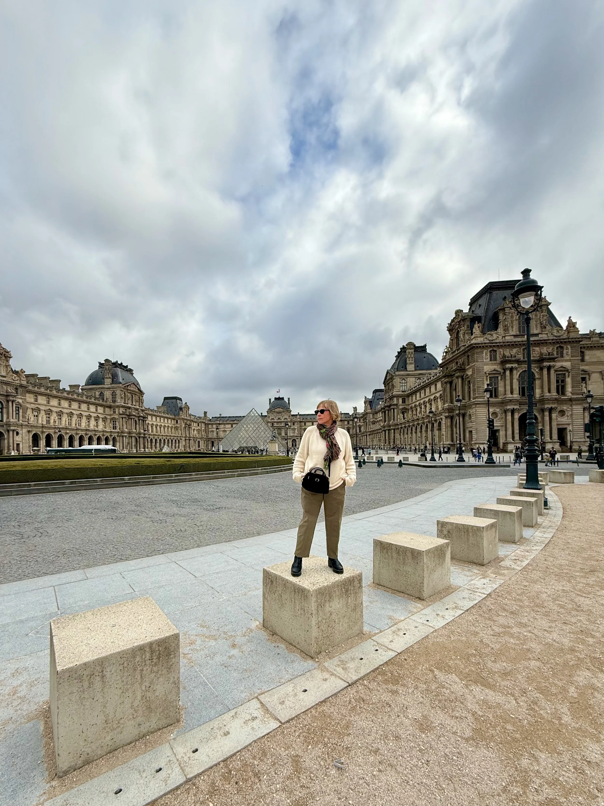 A woman standing on a stone block in a historic European square with ornate buildings and a glass pyramid structure in the background, under a cloudy sky.