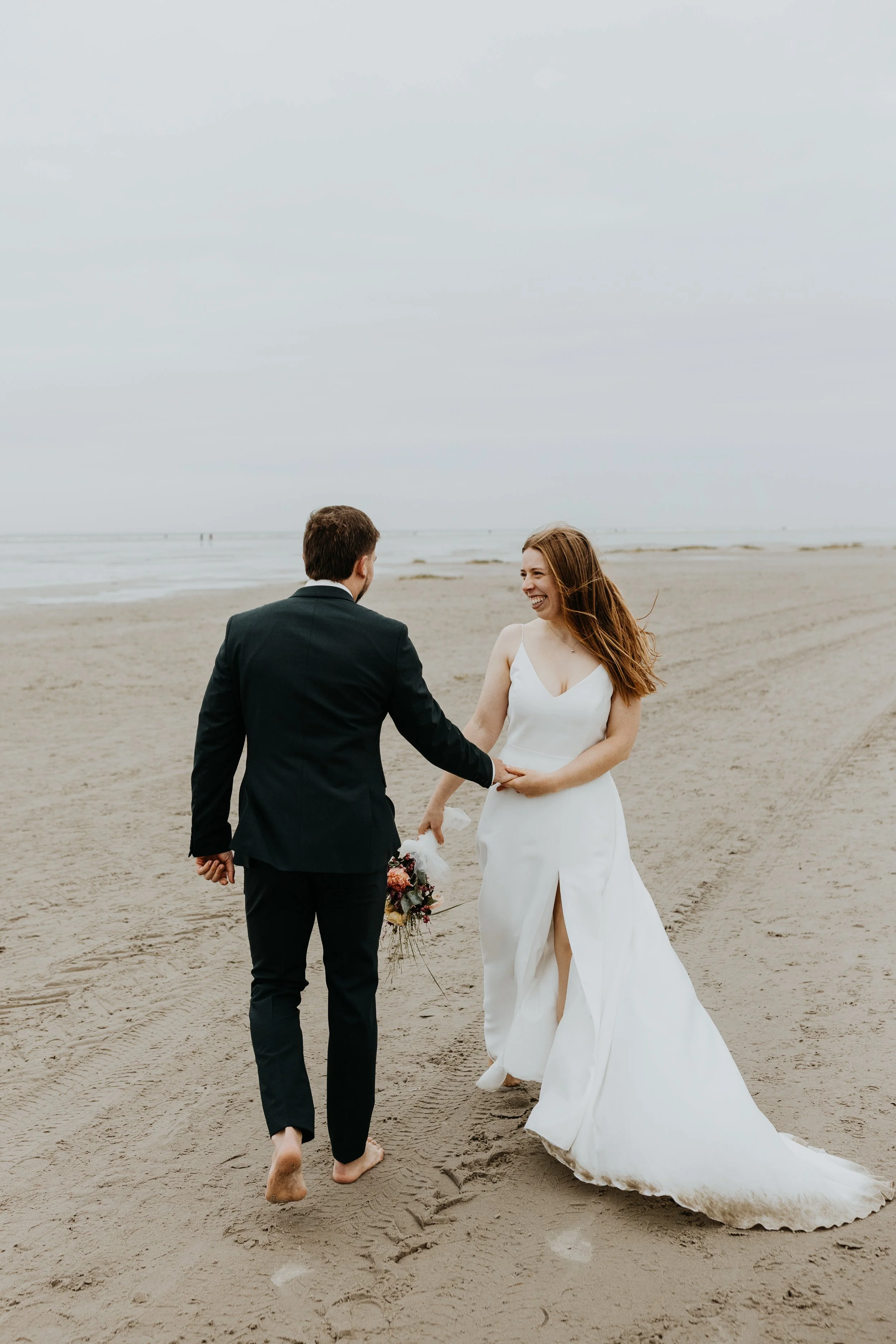 Brautpaar läuft am Strand von Sankt Peter Ording und lacht sich glücklich an. Sie halten sich an den Händen und lassen sich vom Wind treiben.
