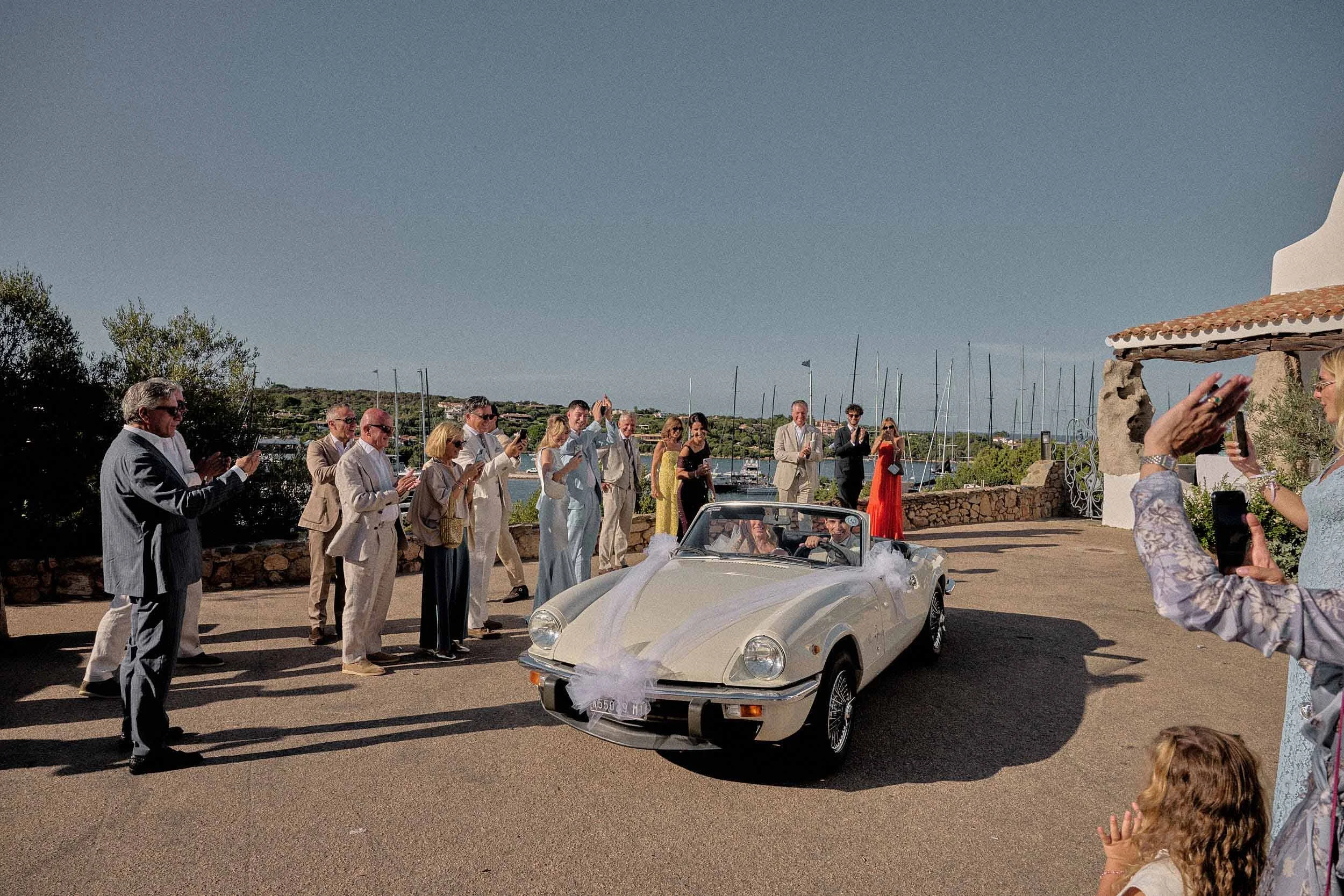 Guests applaud while the newlyweds depart in a decorated classic convertible, with the marina in the background on the Costa Smeralda.