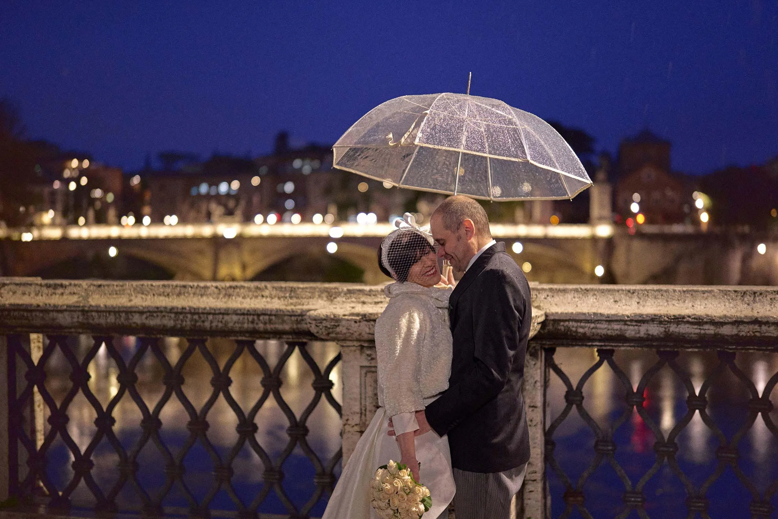 Bride and groom embracing under a transparent umbrella at blue hour, city lights in the background.
