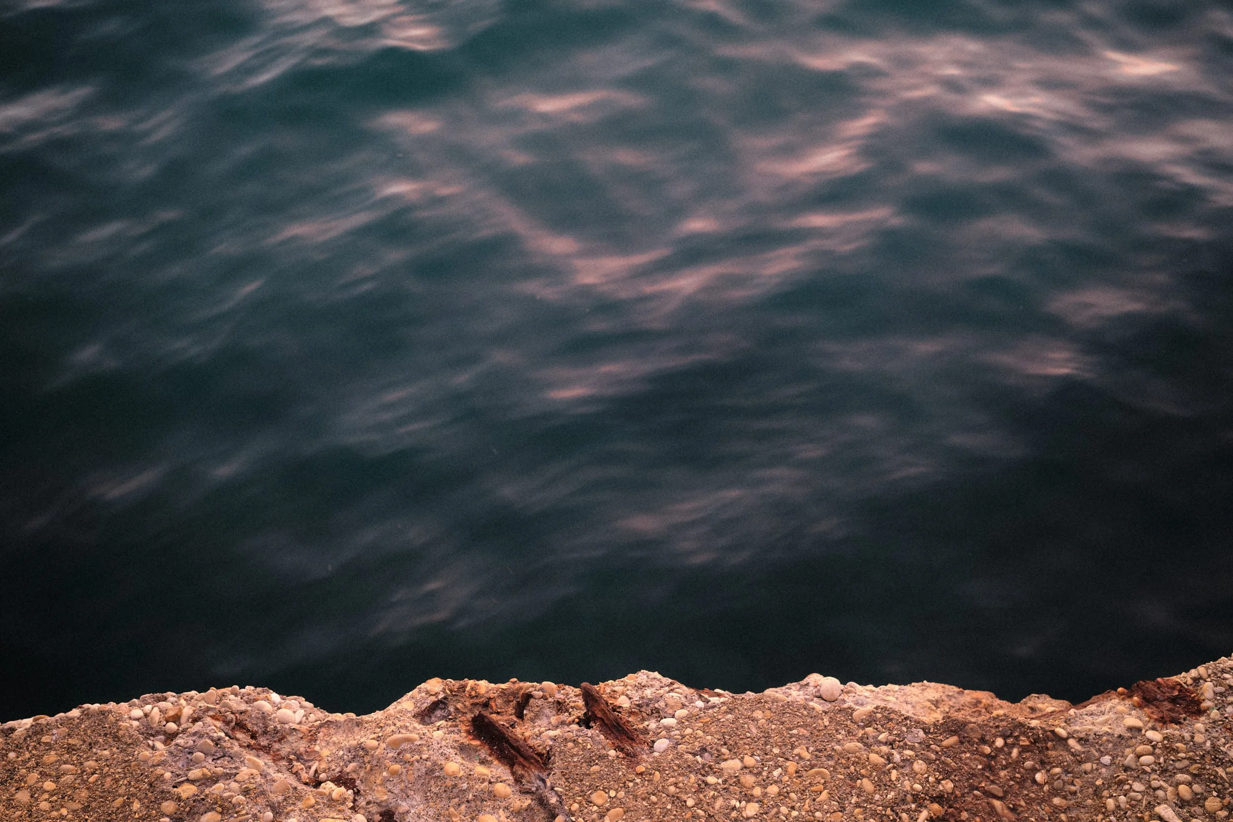 Abstract view of rippling teal water with pink reflections, framed by a rough concrete edge with pebbles and rust in the foreground.