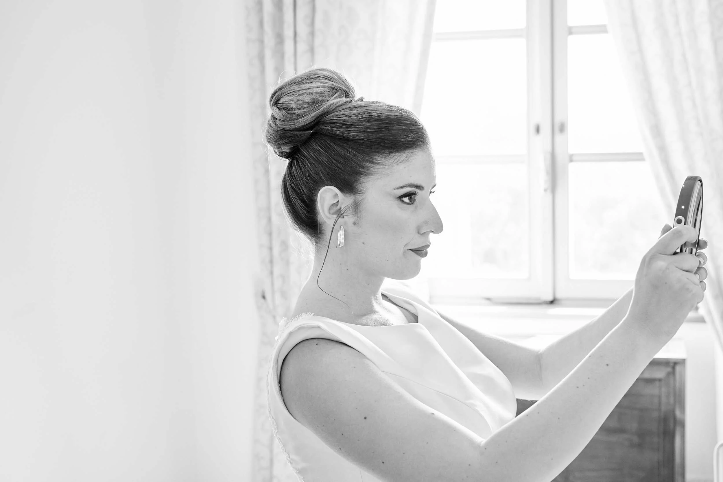 Bride looks into a mirror to check her makeup, a window in the backdrop — black and white.