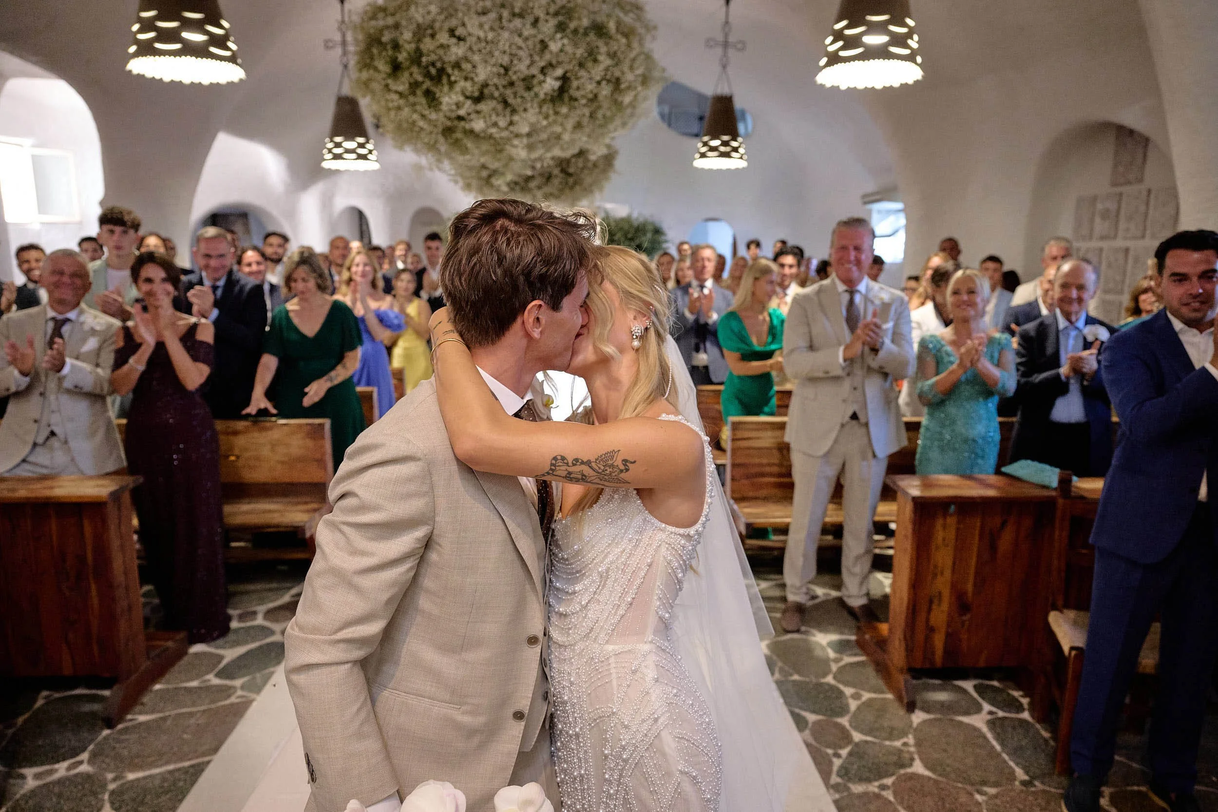 Bride and groom kiss in the aisle as guests applaud inside a whitewashed church in Sardinia.
