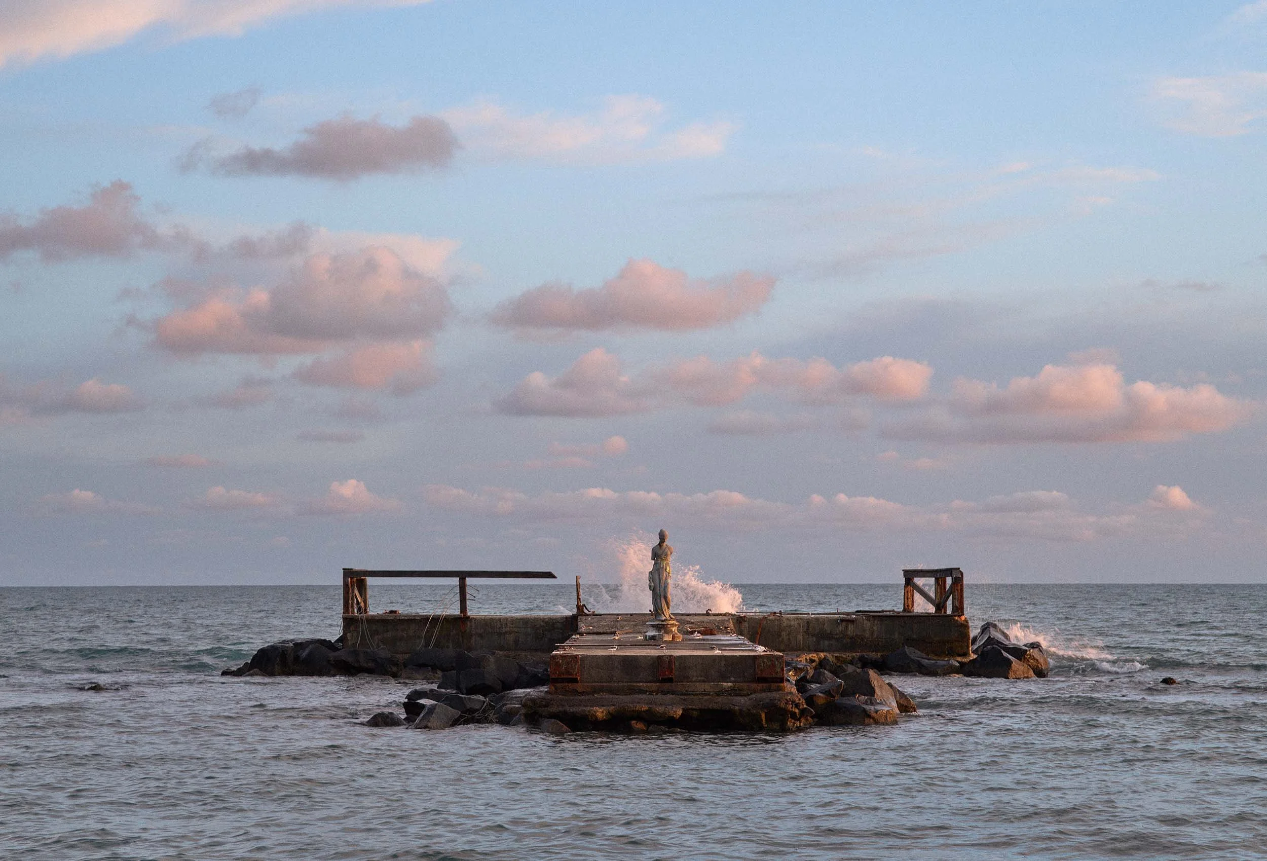 A statue on a concrete pier by the sea, with waves and a wide sky behind.