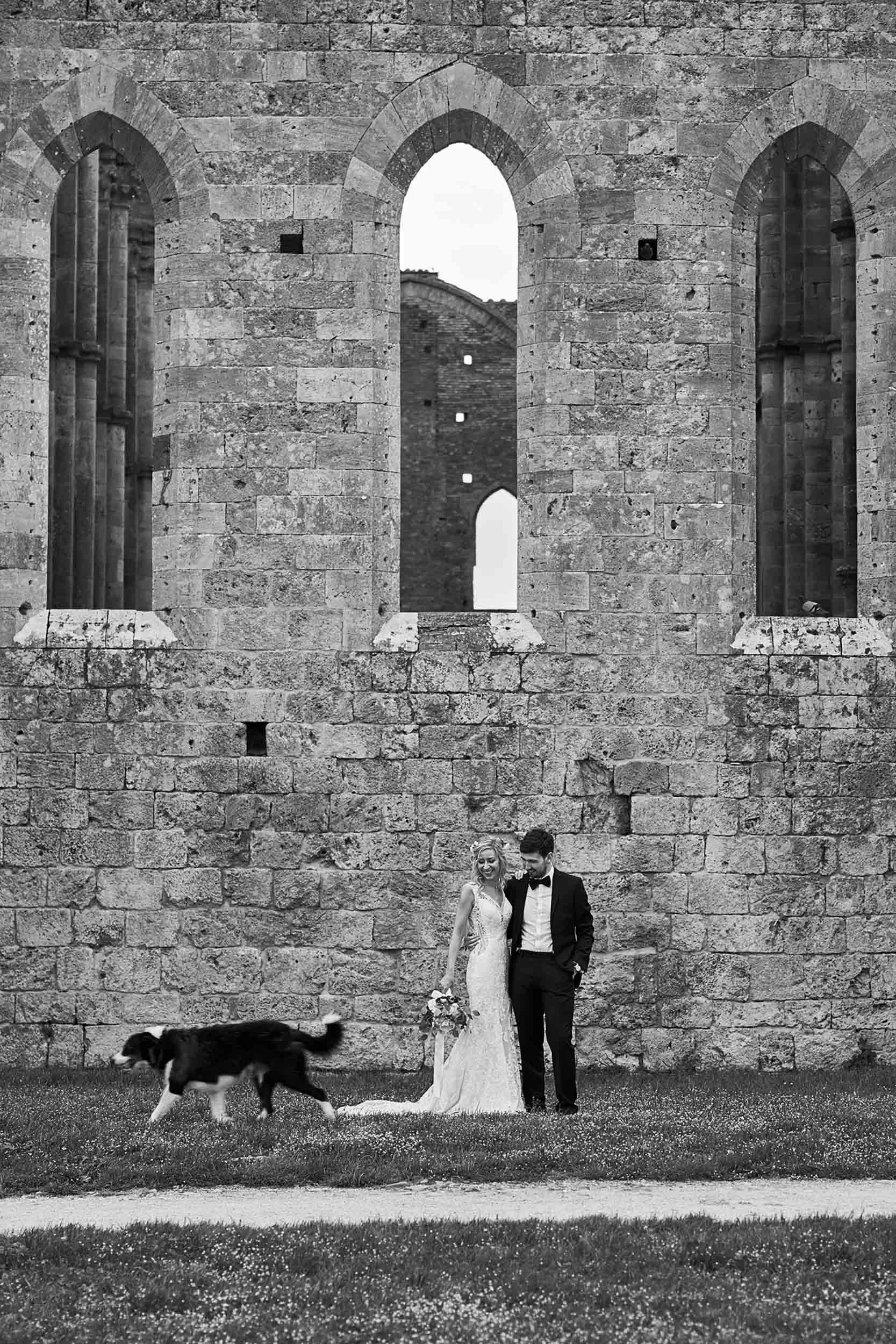 Bride and groom smiling outside the abbey as a dog walks by (black and white)