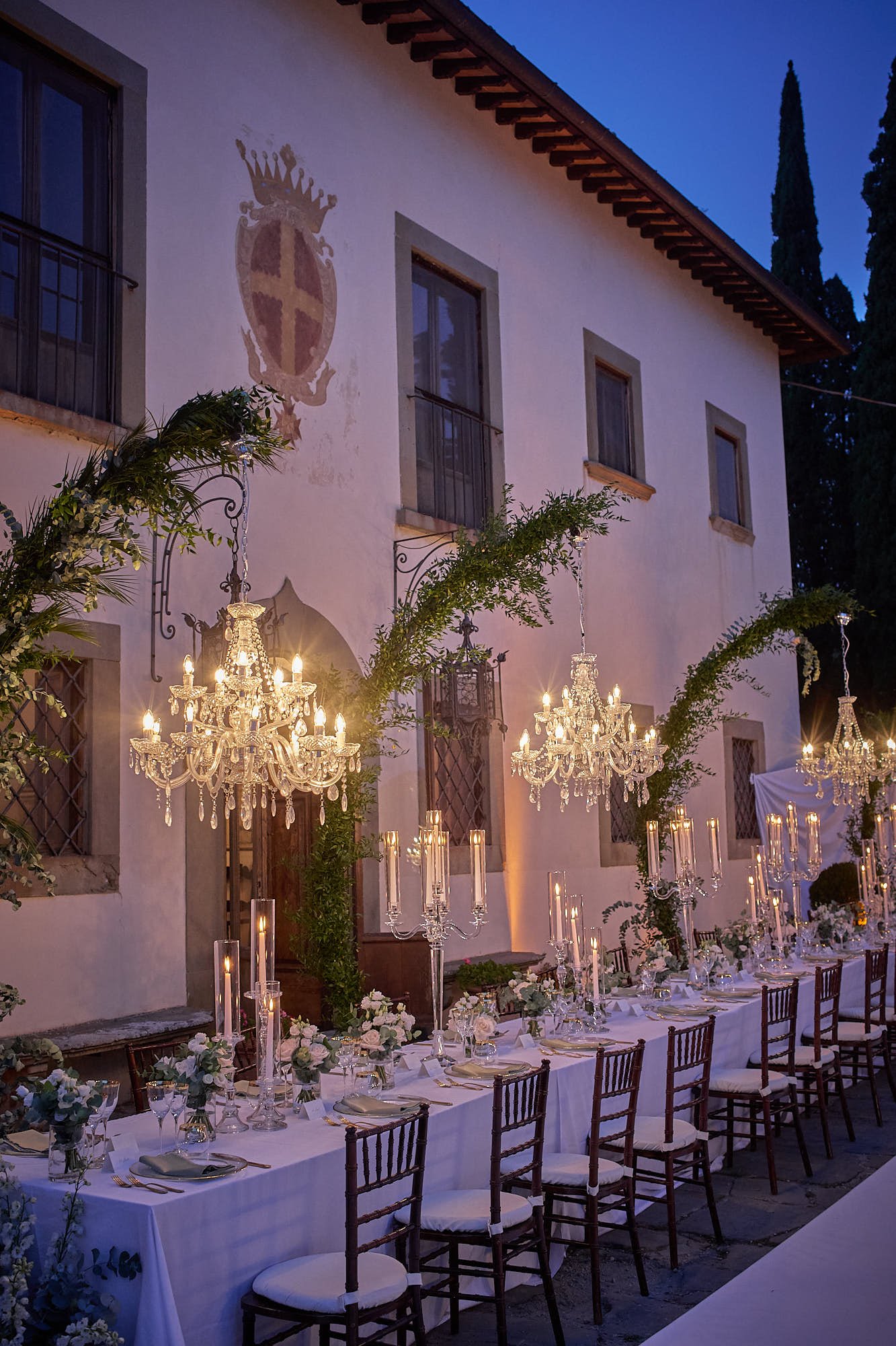 Long outdoor reception table set for dinner with chandeliers, candles, and greenery in a villa courtyard.