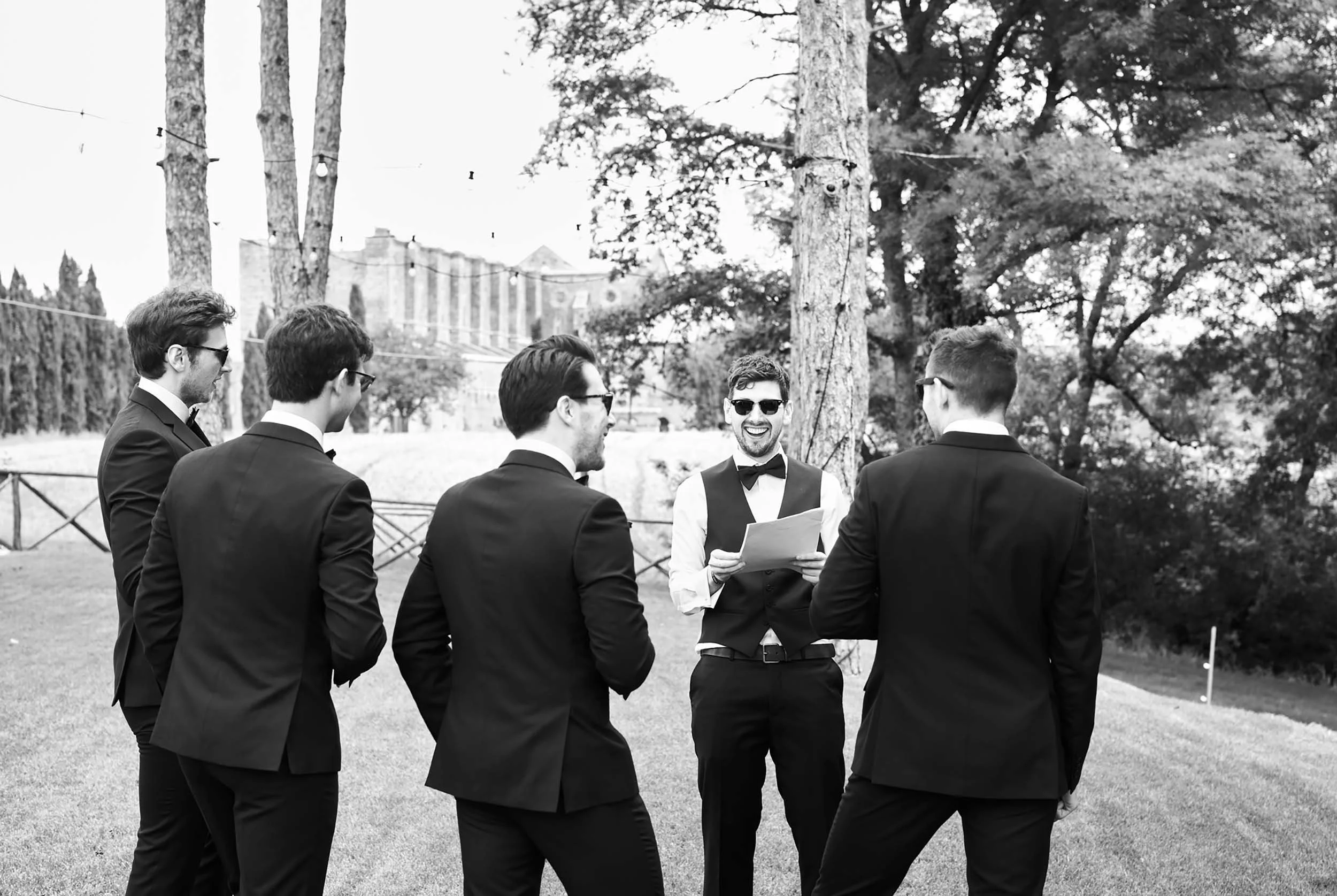 Black and white candid of the groom laughing with groomsmen in front of San Galgano Abbey, Tuscany
