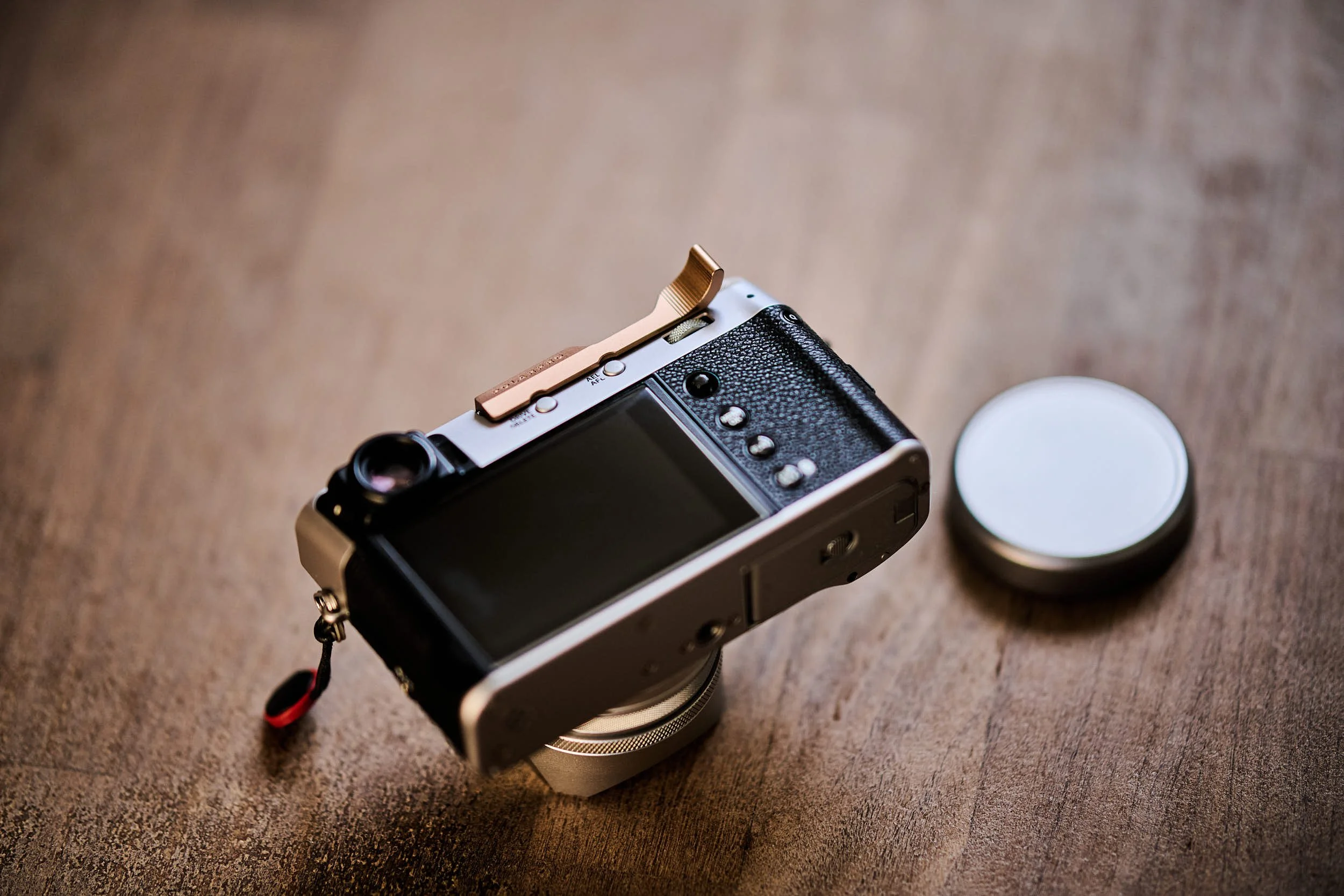Rear view of a silver Fujifilm X100VI balanced on its lens hood, showing the LCD, rear controls and copper PolarPro thumb grip on a wooden table.