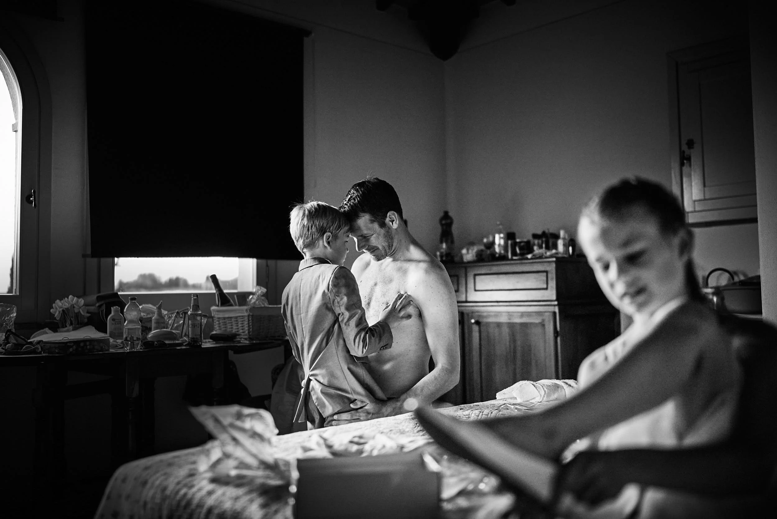 Quiet getting-ready moment: the groom holds a young boy while another child smiles nearby (black and white).