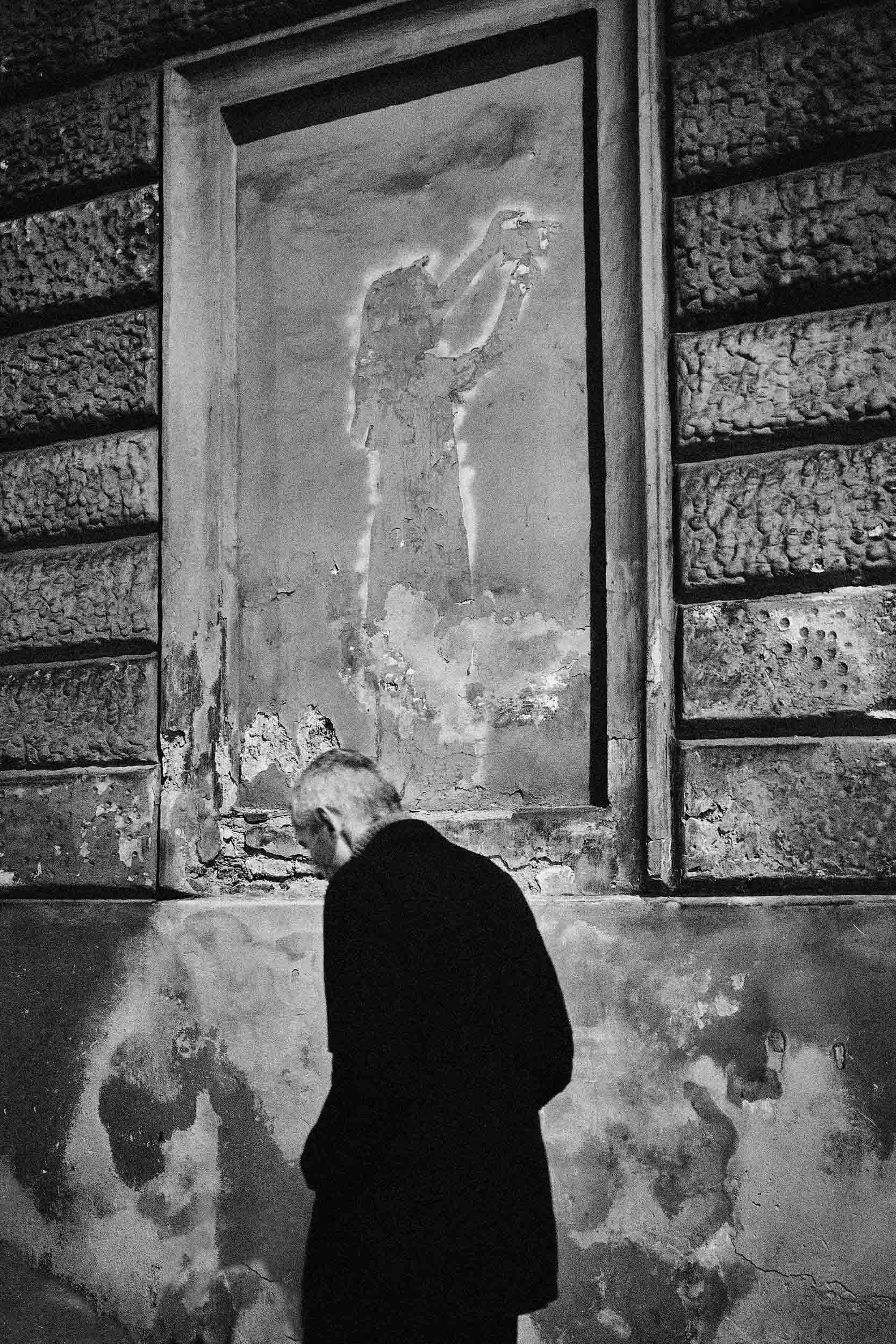 Black and white street photograph in Rome of an elderly man walking past a decayed wall with a faded figure painted holding a camera.