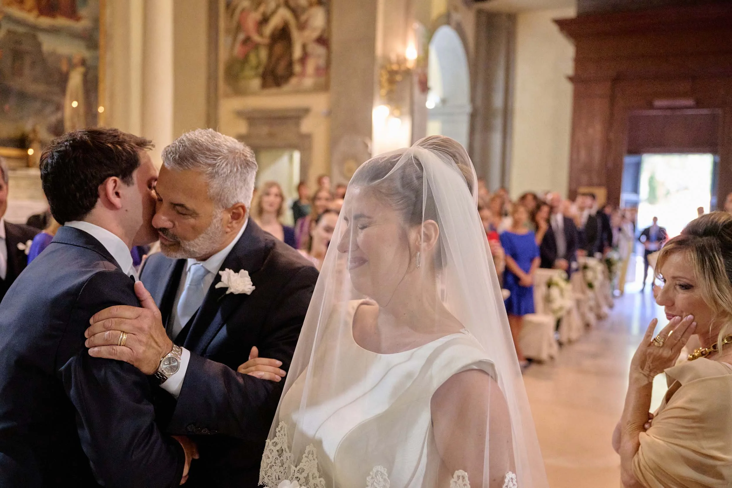 At the altar, the groom is hugged and kissed by the bride’s father; the bride closes her eyes as her mother turns away.