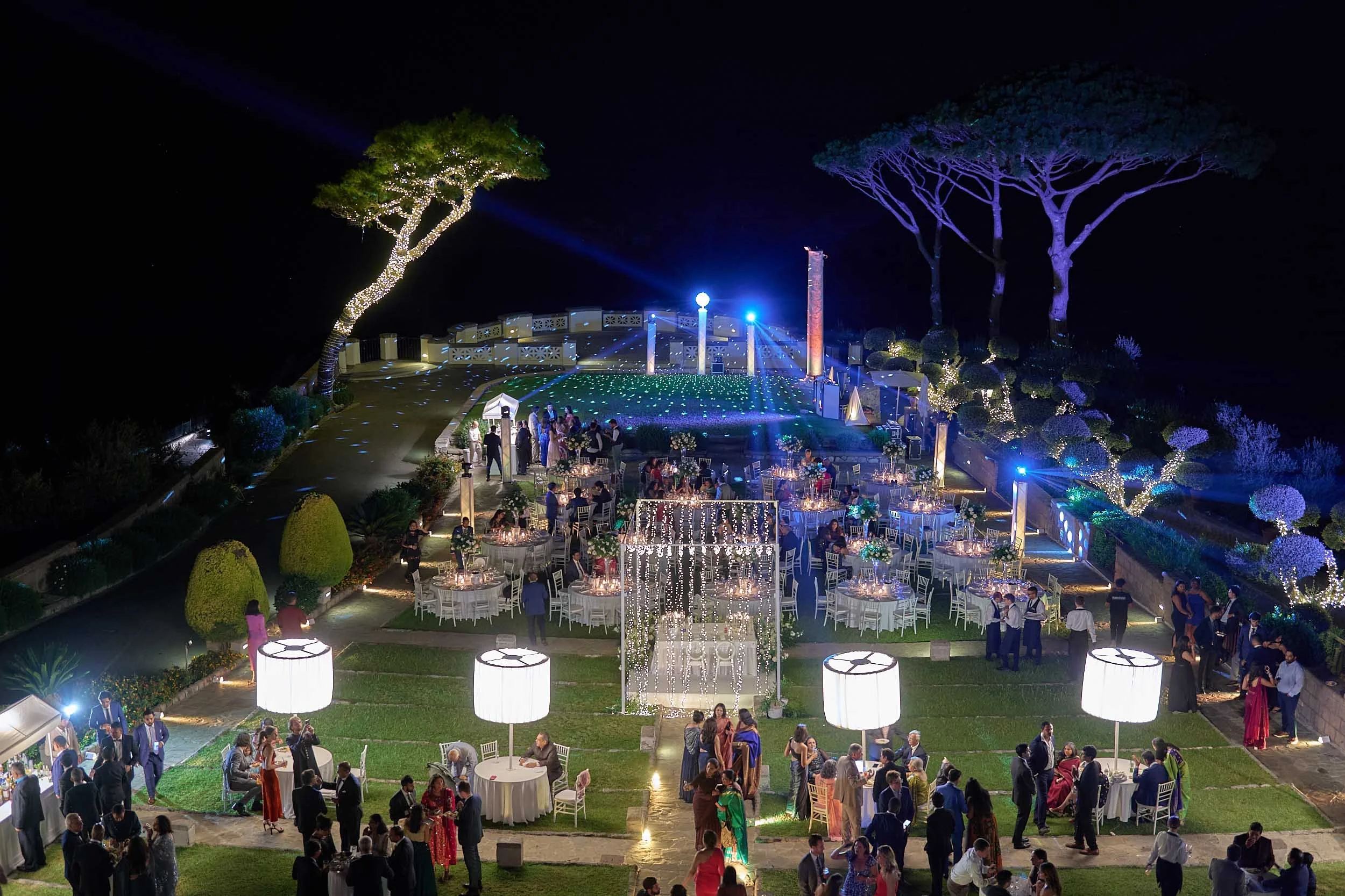 Aerial night view of a garden wedding reception with tables, lights, and guests gathered on a terrace.