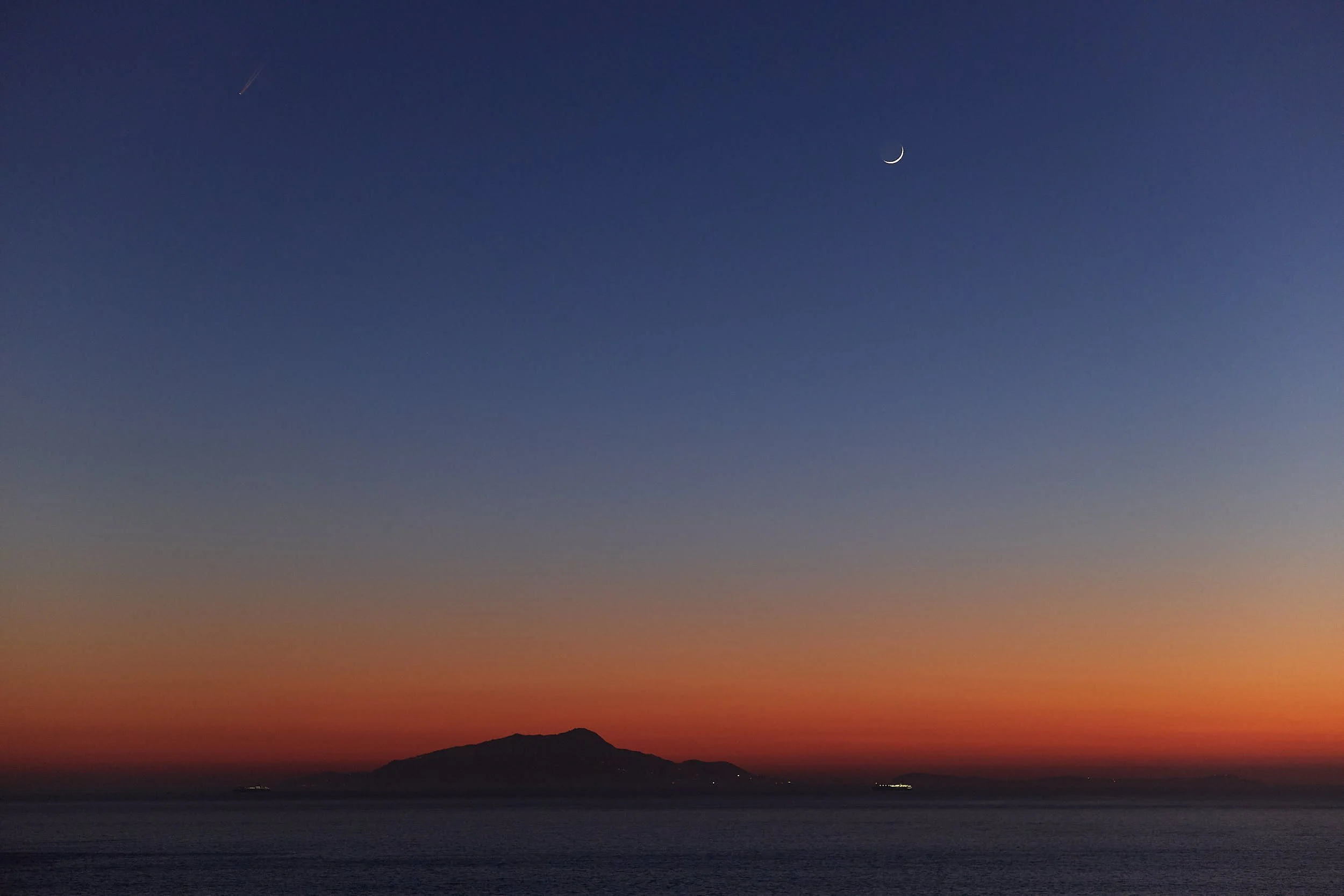 Minimal seascape at dusk with a crescent moon above the horizon and distant coastline silhouette.