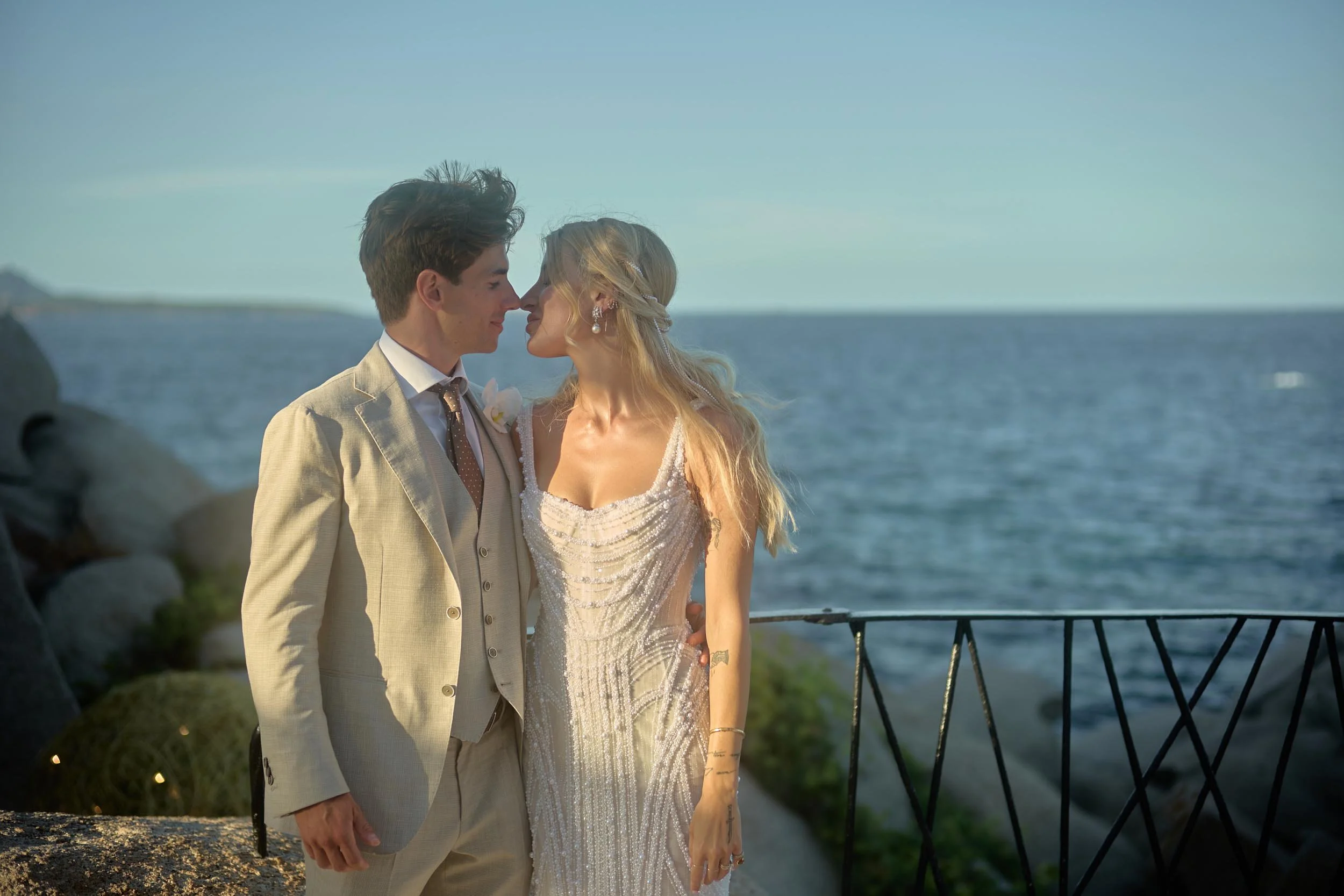 Bride in a beaded gown and groom in a light suit leaning in, almost kissing at golden hour, with the sea in the background in Sardinia.