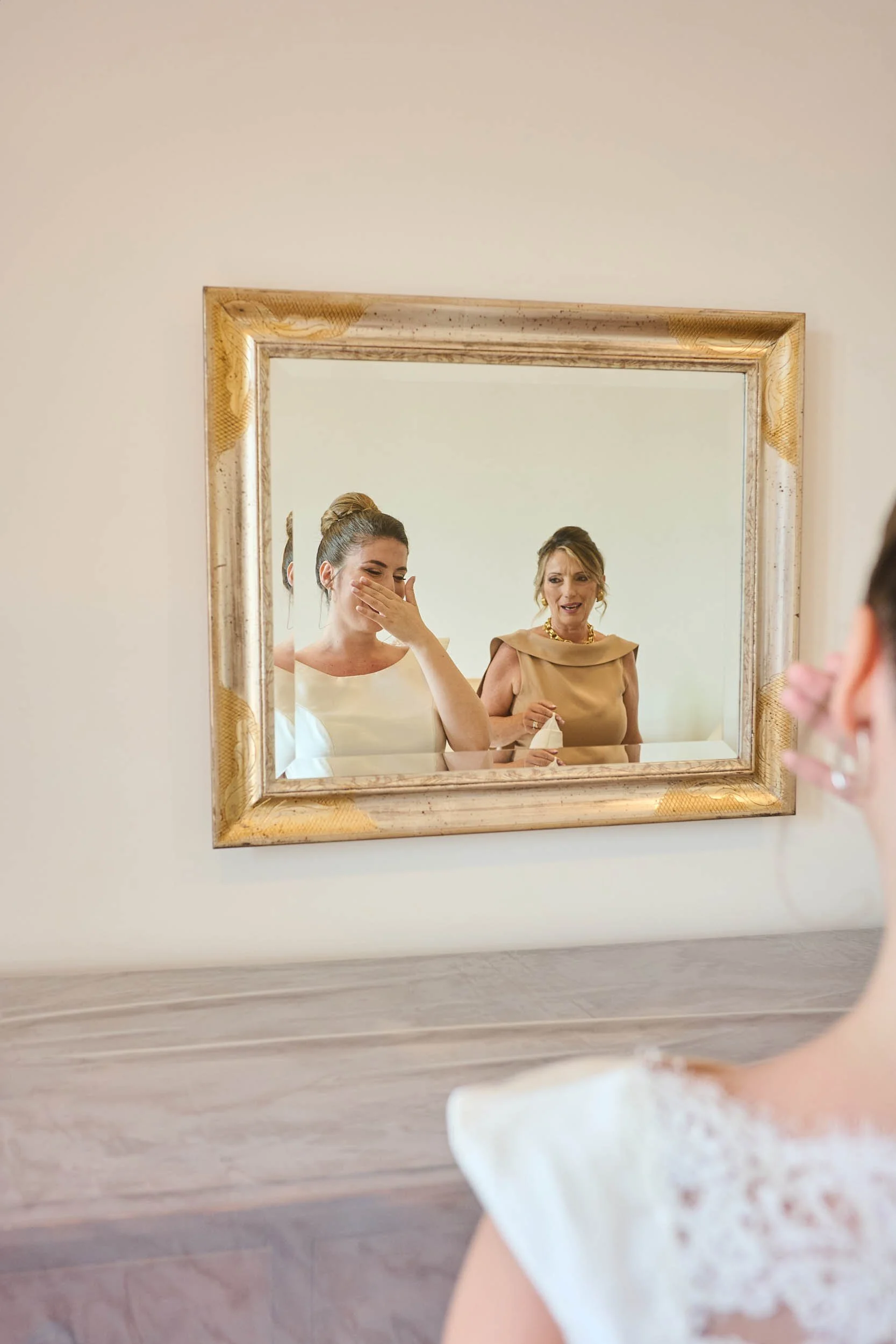 Emotional bride in front of a mirror, covering her mouth as she smiles while her mother looks on.