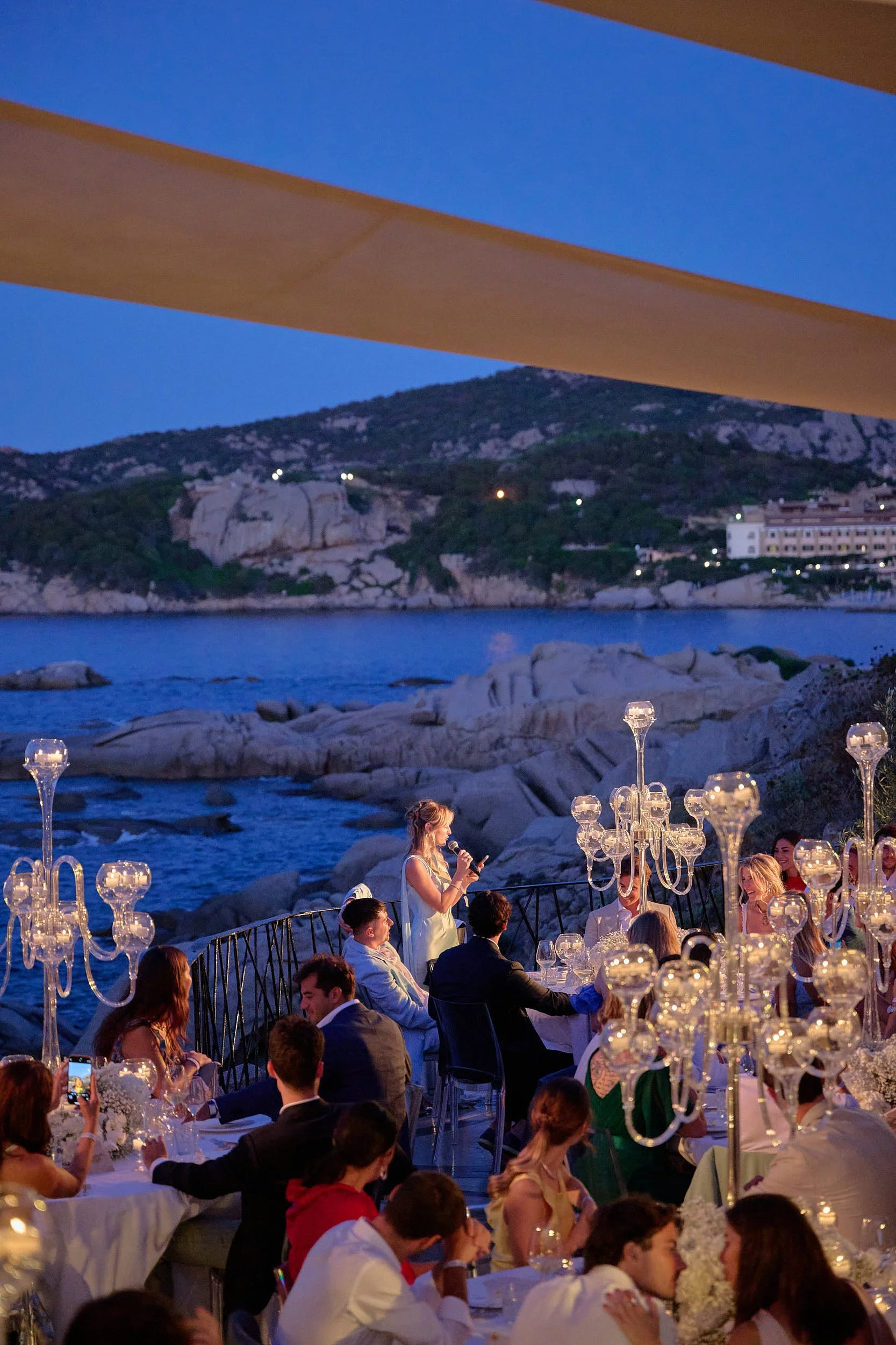 Wide reception dinner scene with a speech in progress, glass candelabras and the rocky coastline in the background, Sardinia (Costa Smeralda).