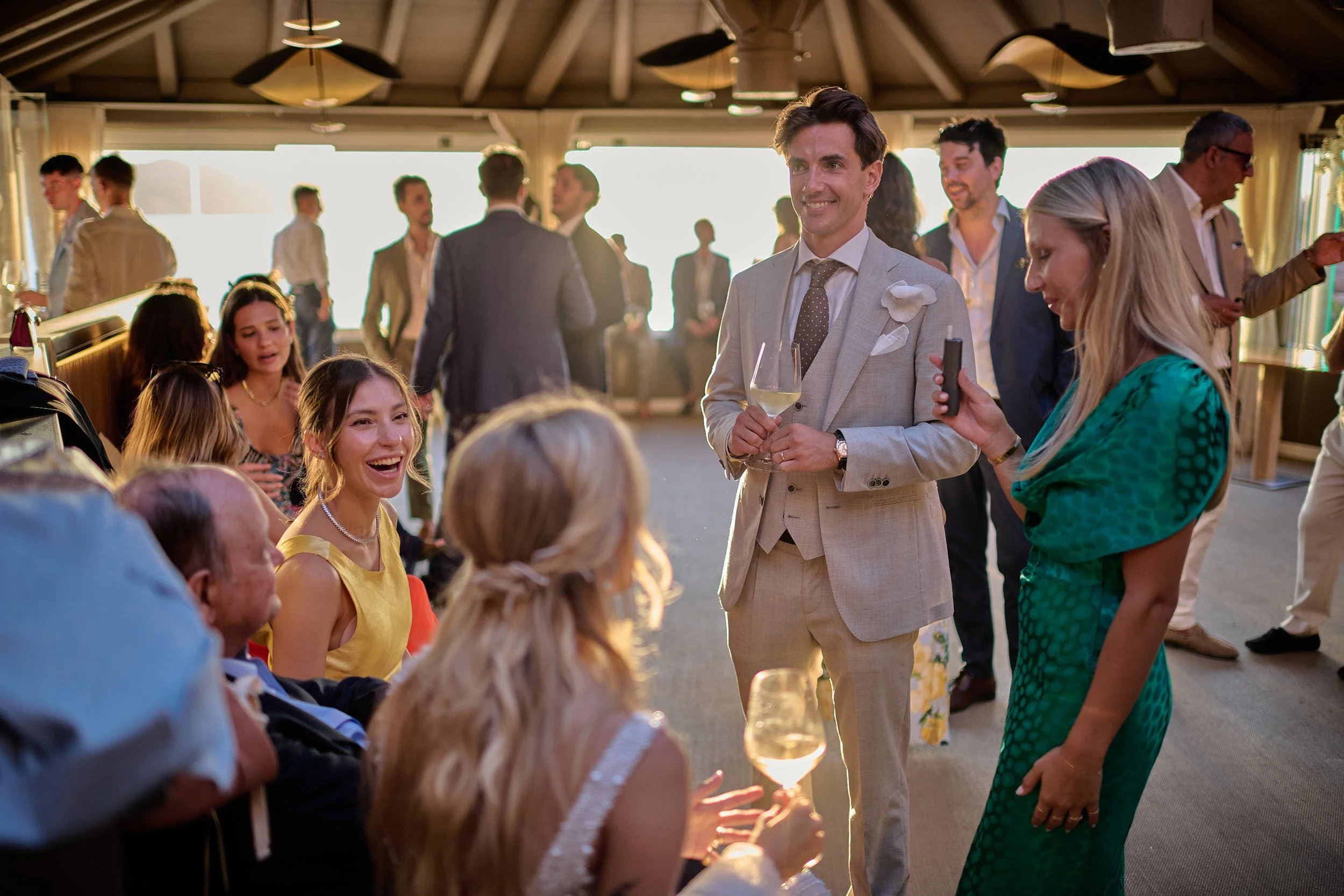 Groom in a light suit smiling during cocktail hour with guests, backlit by warm sunset light at a seaside wedding in Sardinia.