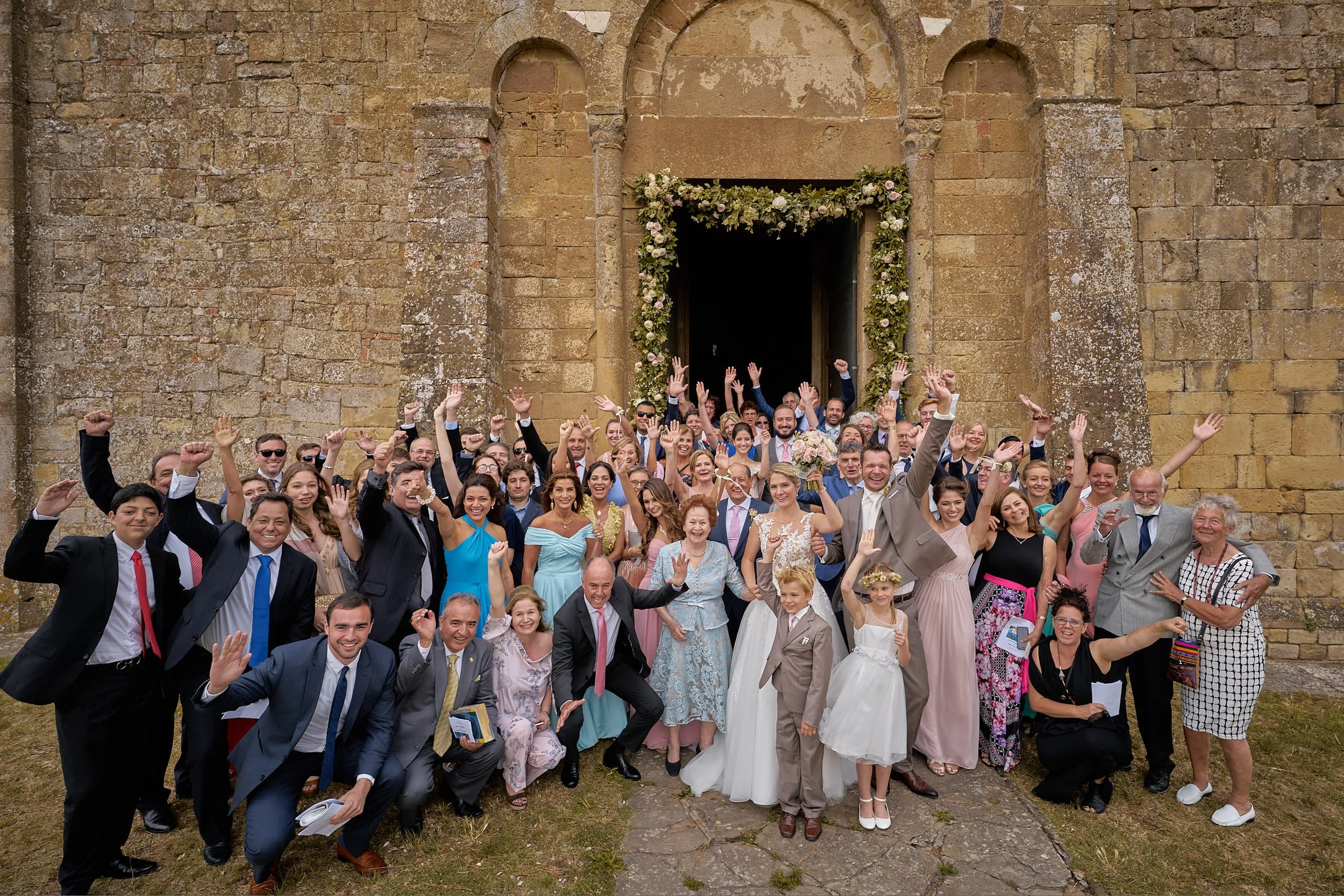 Large group celebrating outside a historic stone church, hands raised, with the couple centered near the entrance