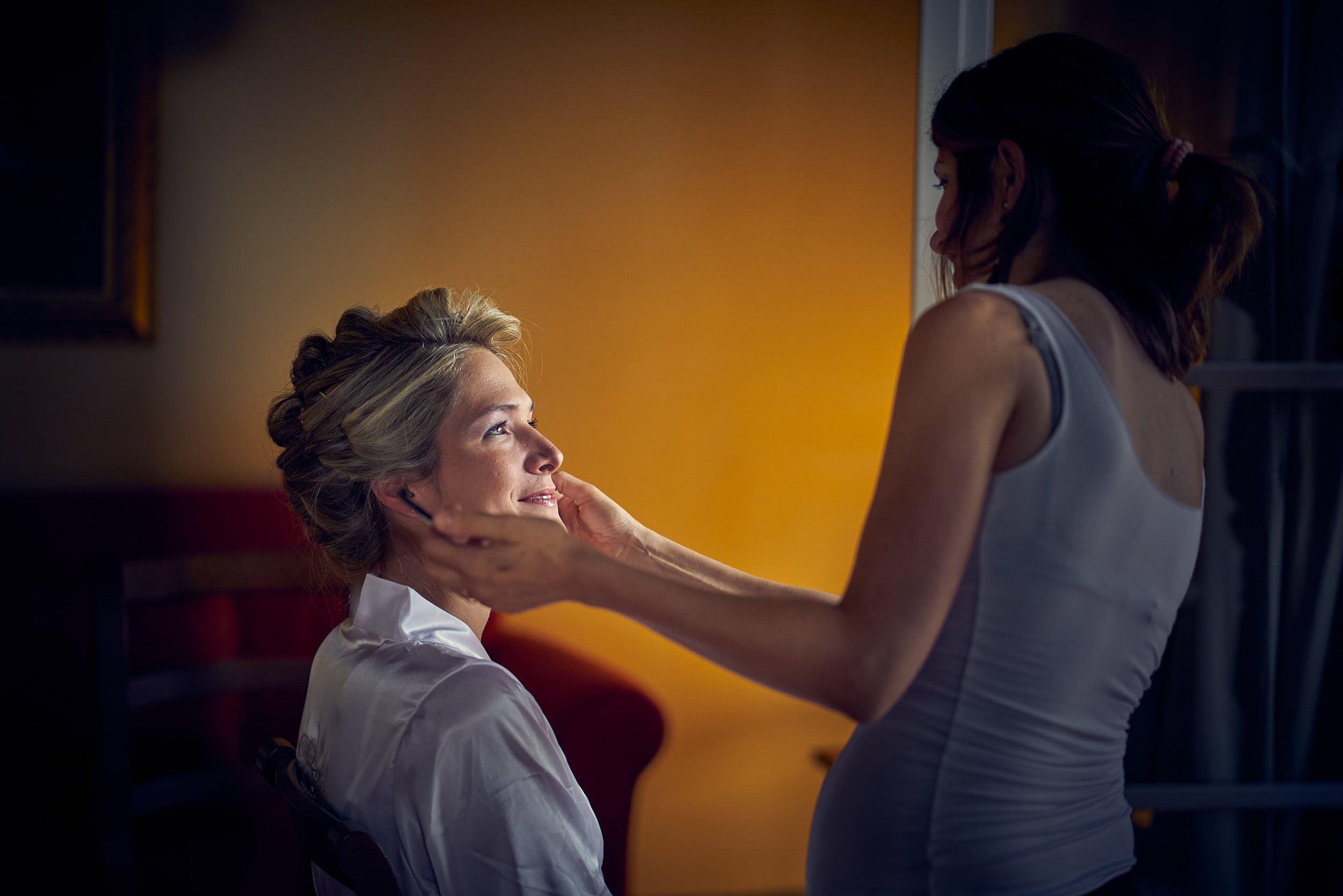 Bride in a robe during preparations as the makeup artist adjusts her face in warm window light.