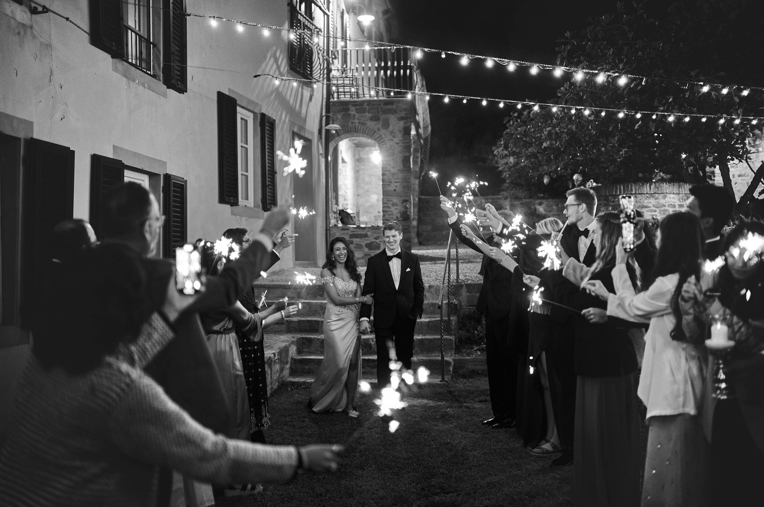 Couple arrives at the wedding dinner surrounded by guests and sparklers — black and white, Destination Wedding Tuscany