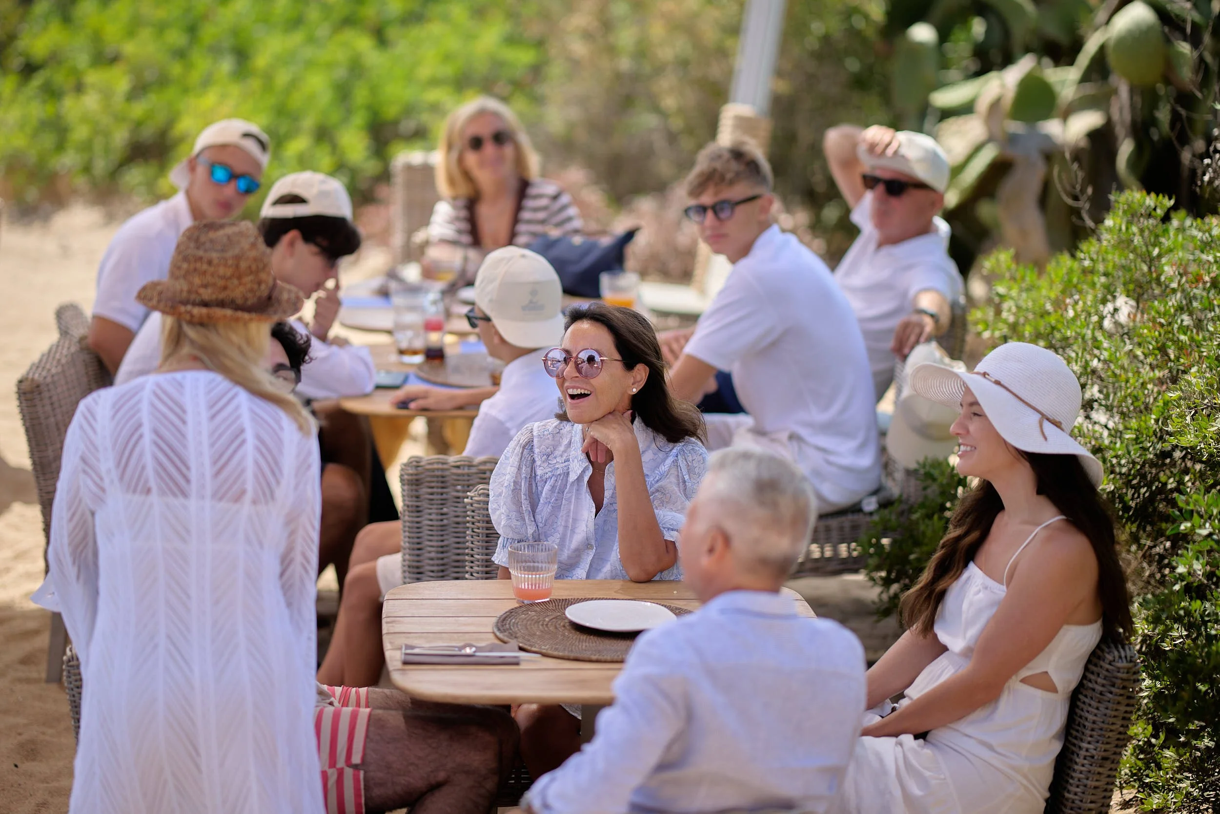 Candid moment at a day-after beach brunch: guests gathered around a table in summer hats and sunglasses, laughing in the shade on the Costa Smeralda.