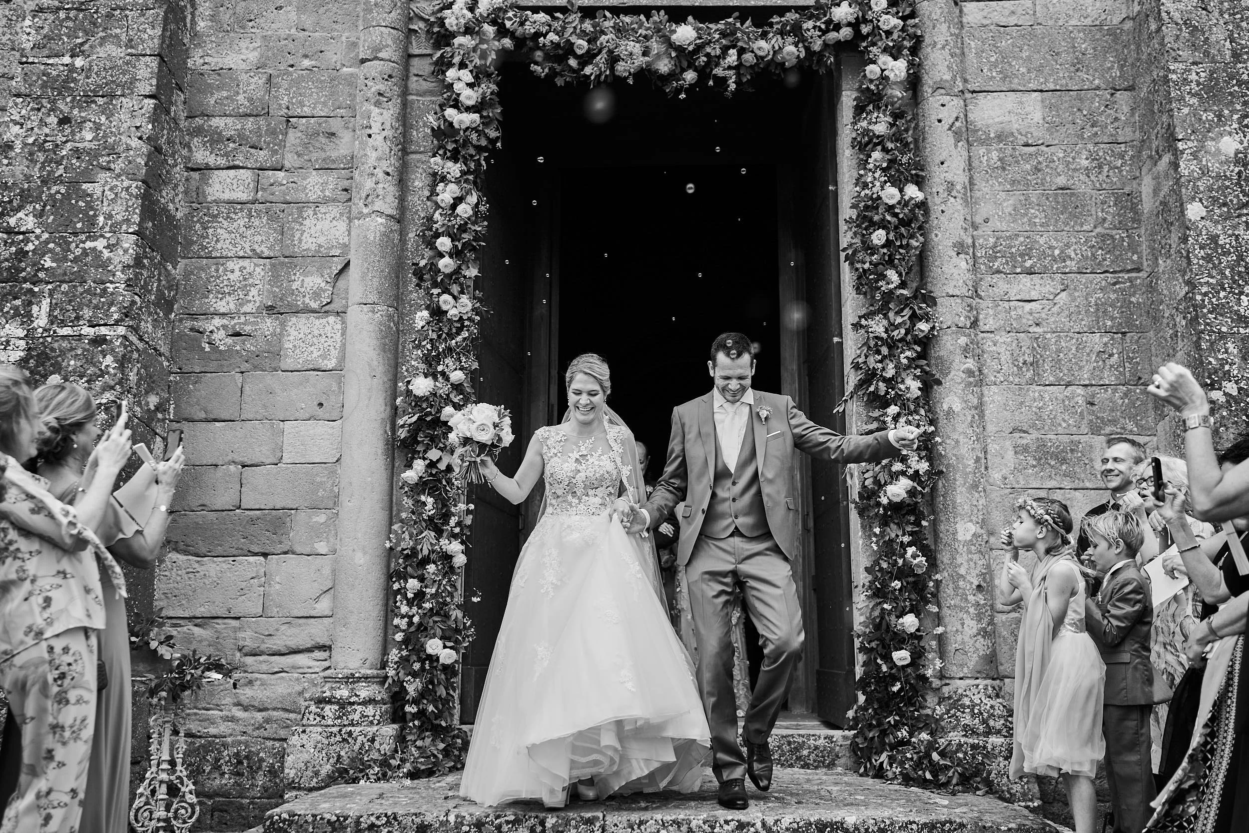 Black-and-white church exit with the couple framed by a lush floral arch and guests on both sides, a timeless documentary moment.