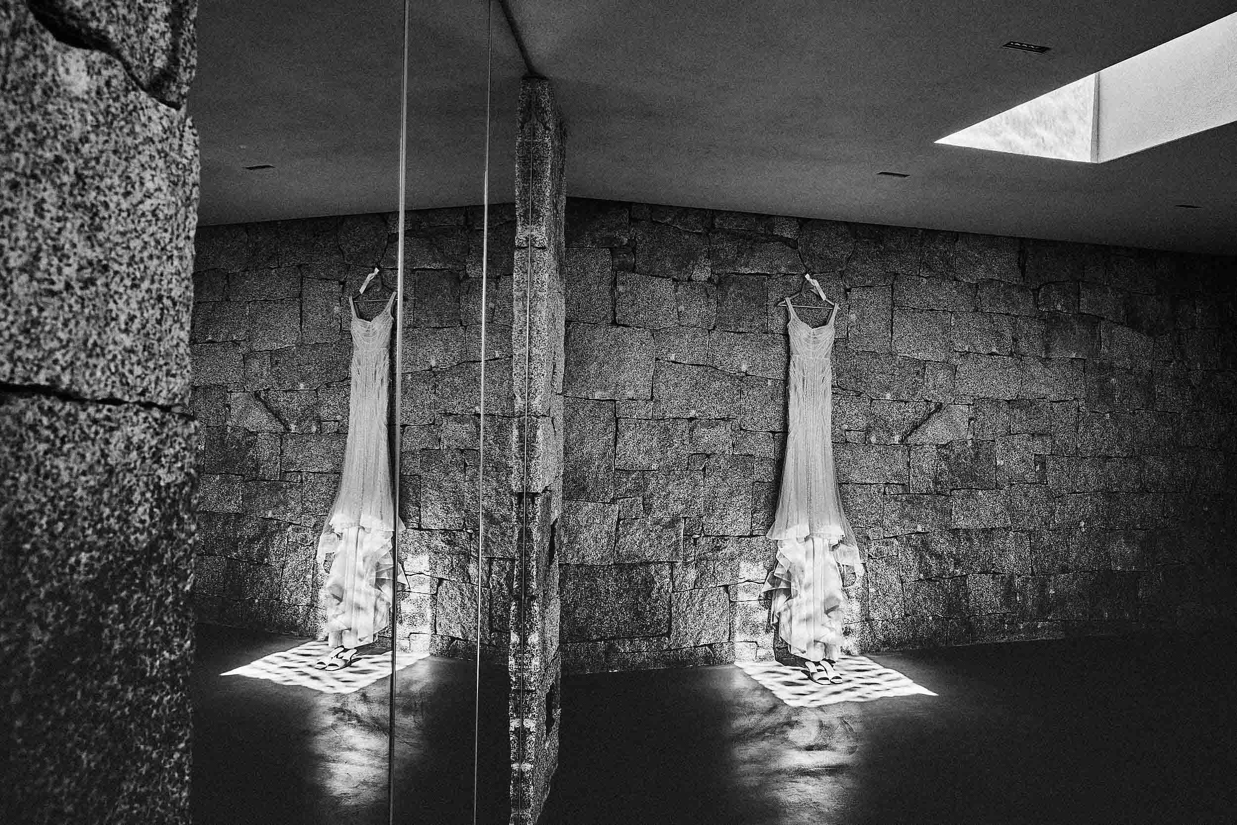 Wedding dress reflected in glass, hanging in a stone-walled room in Sardinia.