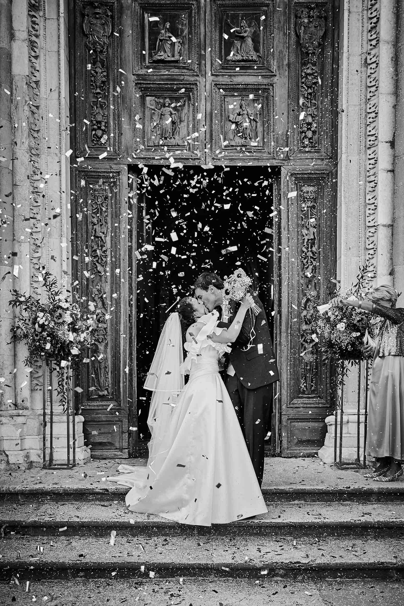 Bride and groom kissing as they exit the church under a shower of confetti, black and white documentary wedding photography in Todi.