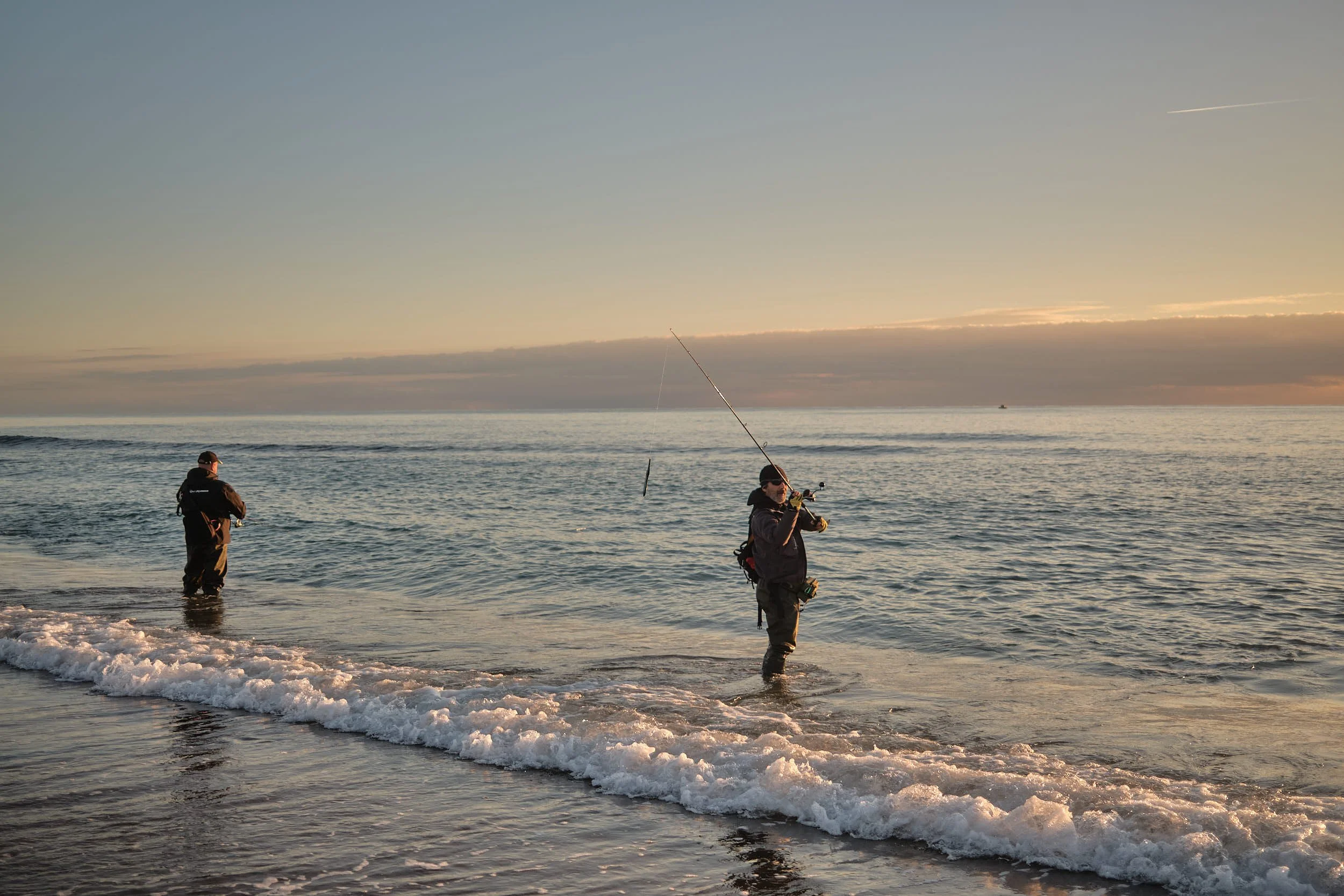Two anglers standing in the surf at sunrise, one casting a fishing line as soft warm light reflects on the sea.