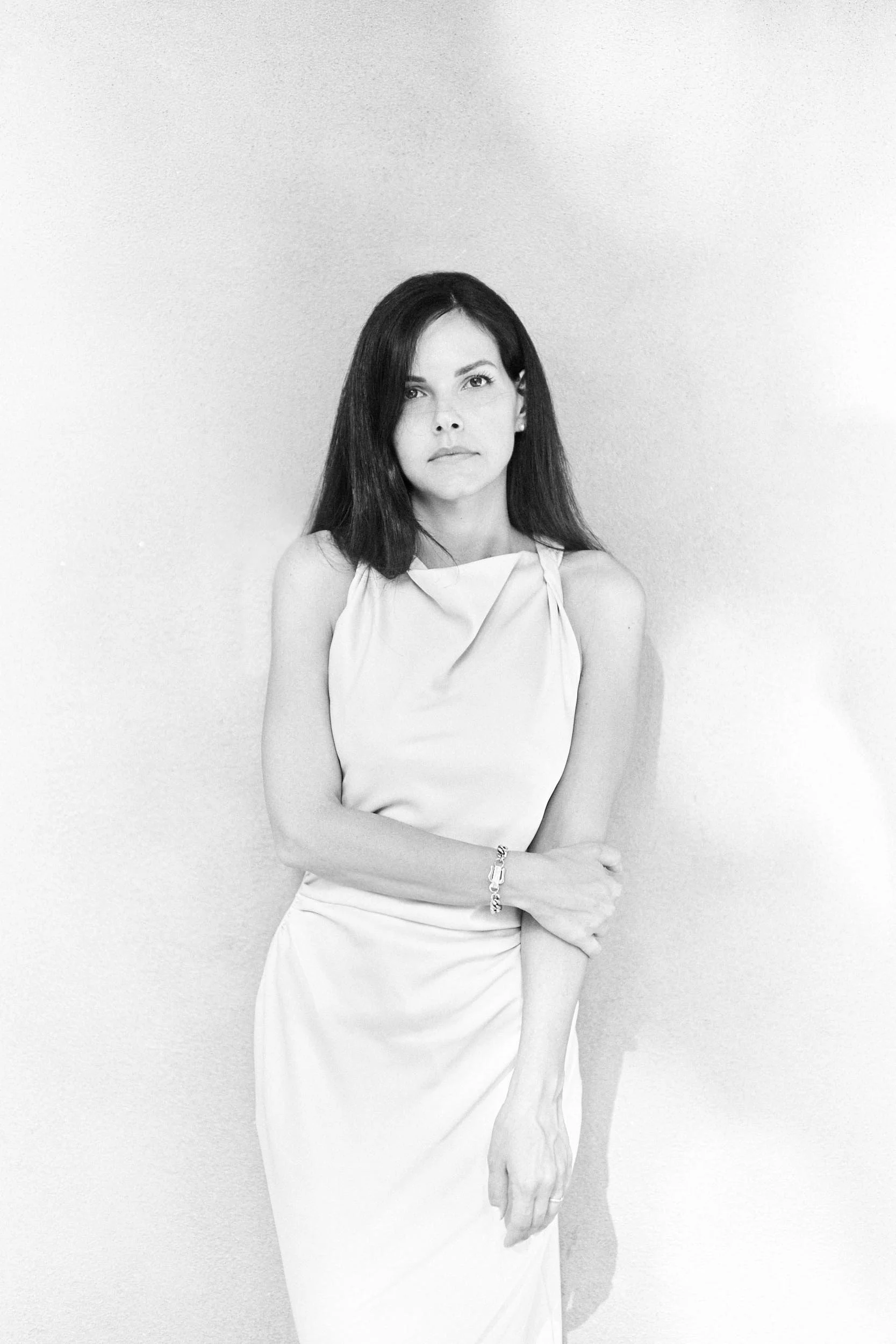 Black-and-white portrait of a woman in a light dress against a plain wall, looking into the camera.