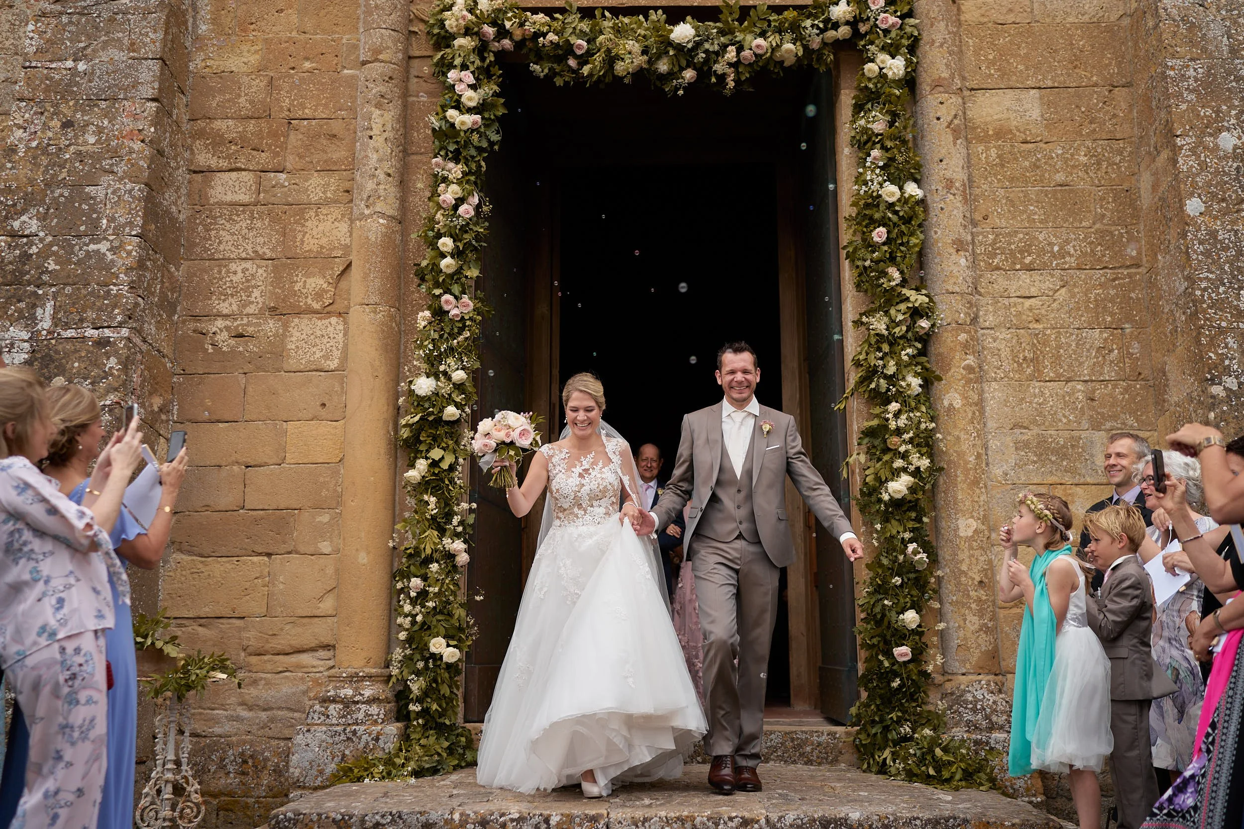 Bride and groom walk out of a stone church under a floral arch as guests celebrate outside.