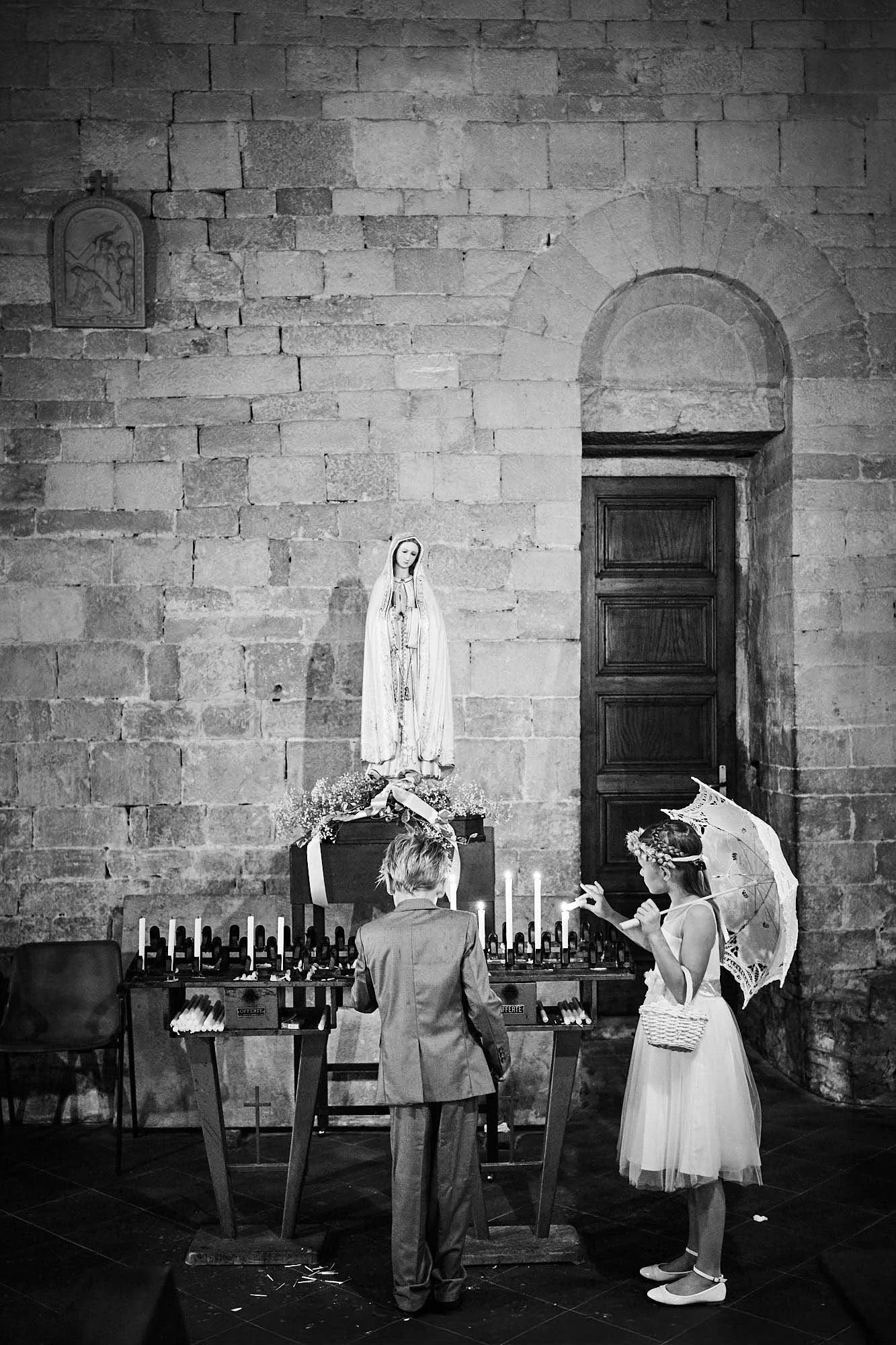Two children lighting votive candles beneath a statue in a stone church, a quiet wedding moment