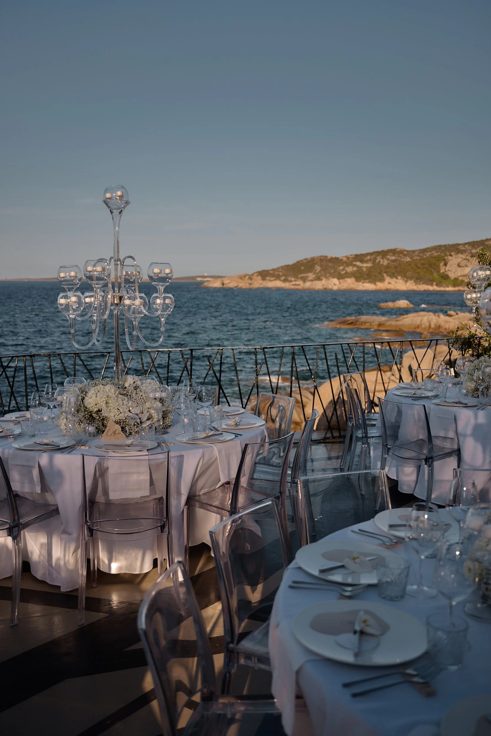 Wedding reception tables with white linens, clear chairs and glass candelabras overlooking the sea at golden hour in Sardinia (Costa Smeralda).