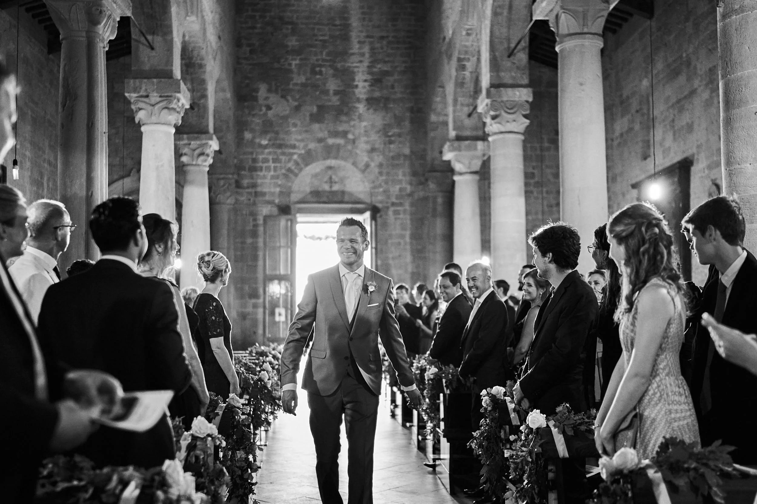 Groom walking toward the ceremony in a stone church, smiling as guests watch from both sides