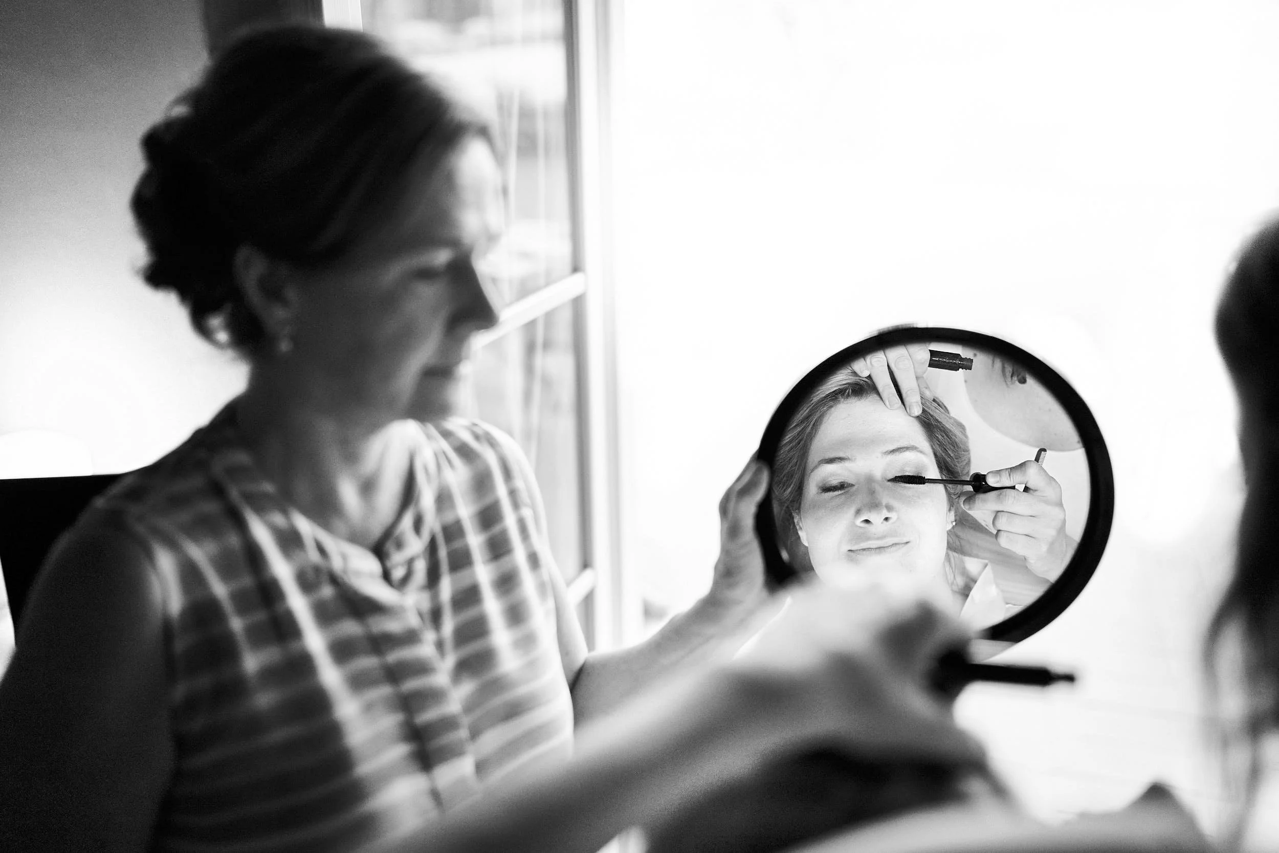 Makeup artist applying mascara as the bride closes her eyes, reflected in a round mirror with soft window light