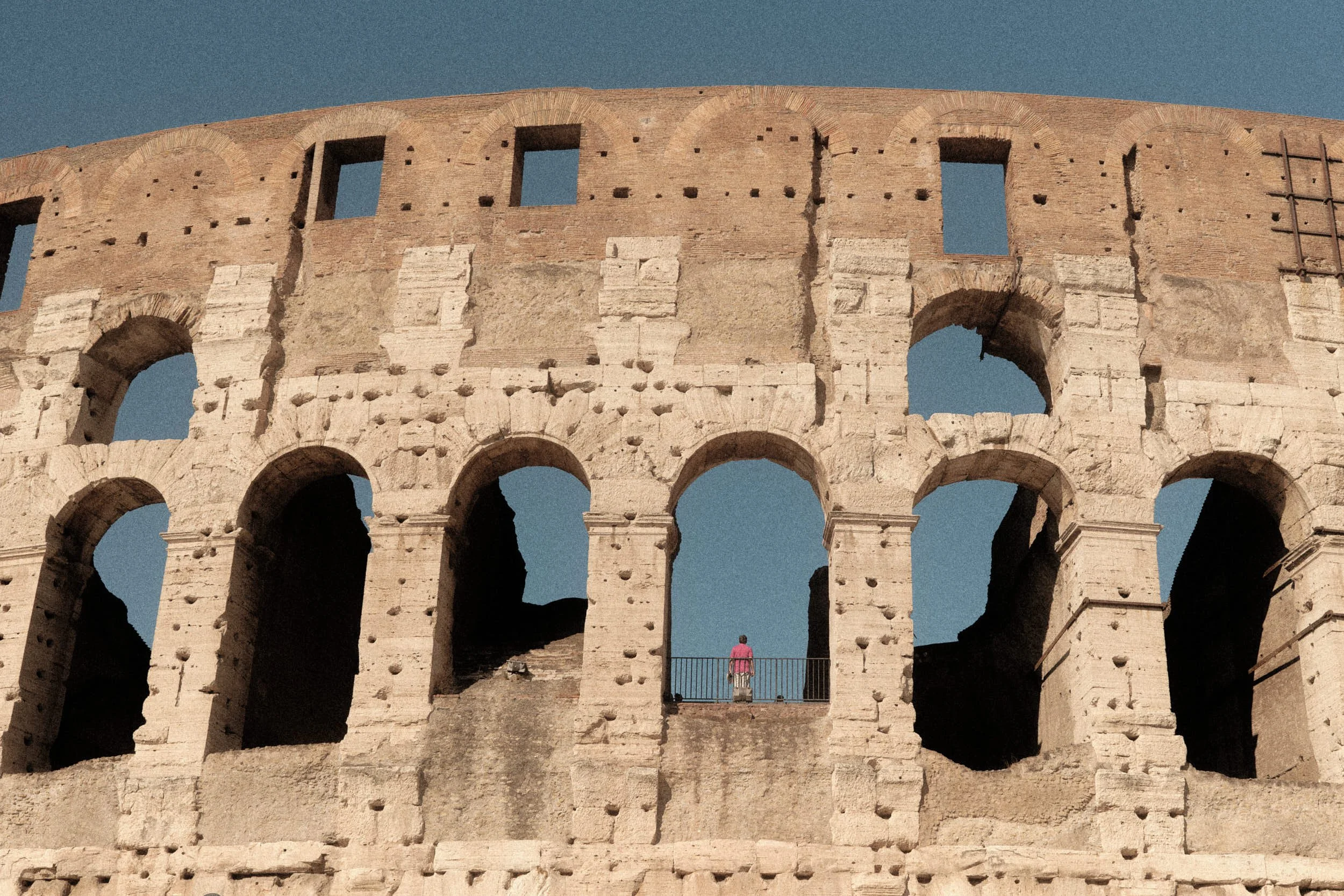 Detail of the Colosseum arches in Rome with a single person in a pink shirt standing on a balcony against a blue sky.