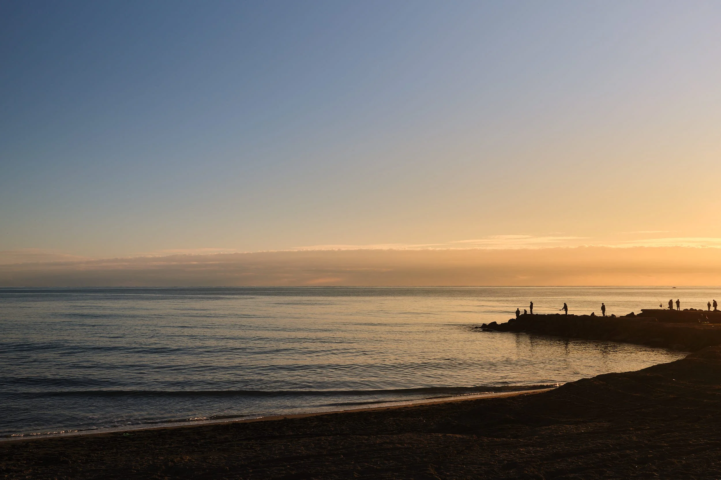 Wide seascape at sunset with calm waves, golden light on the horizon and small silhouettes of people standing on a rocky pier.