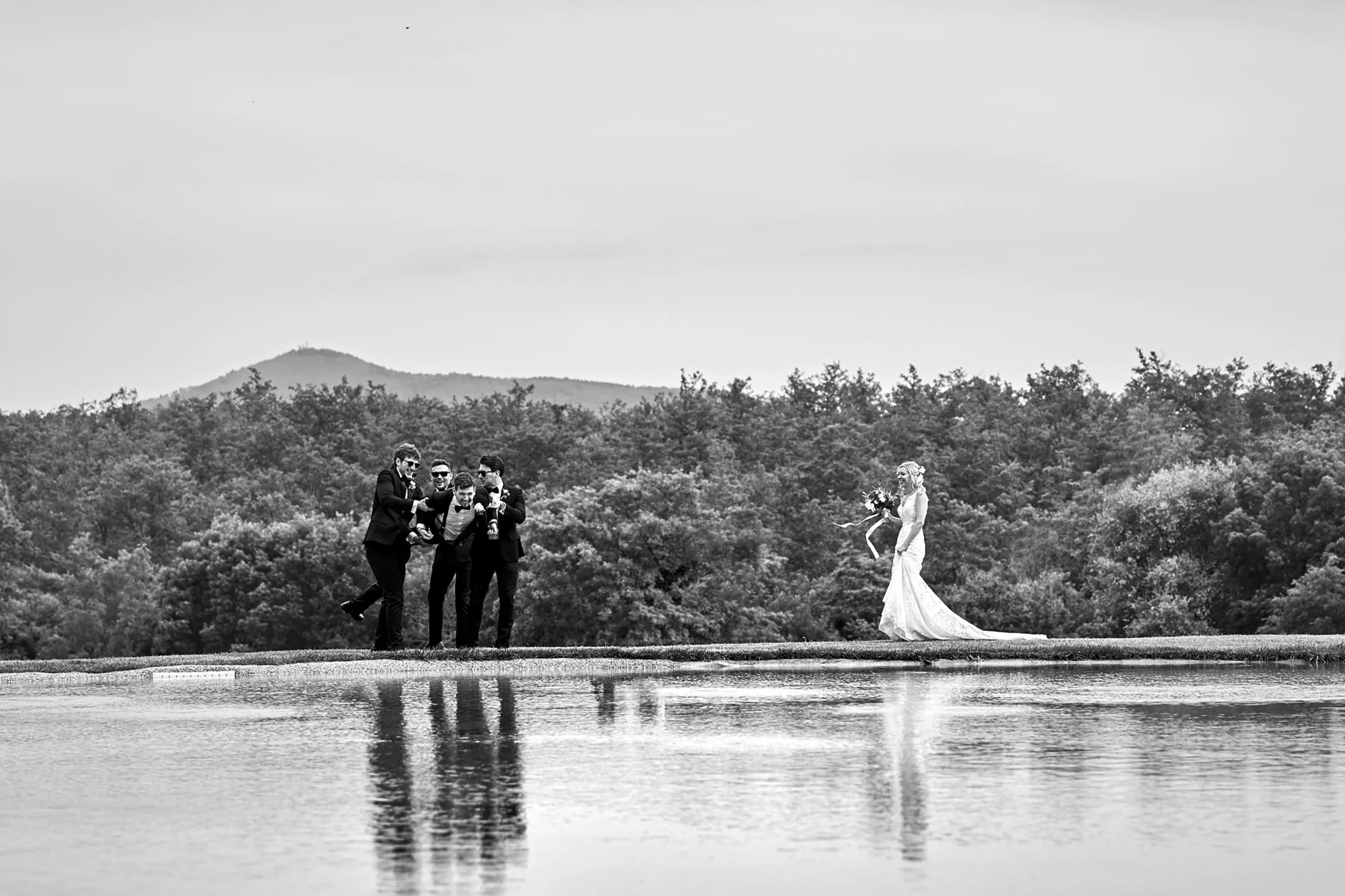 Groomsmen jokingly lift the groom near a pond in front of the bride (black and white)
