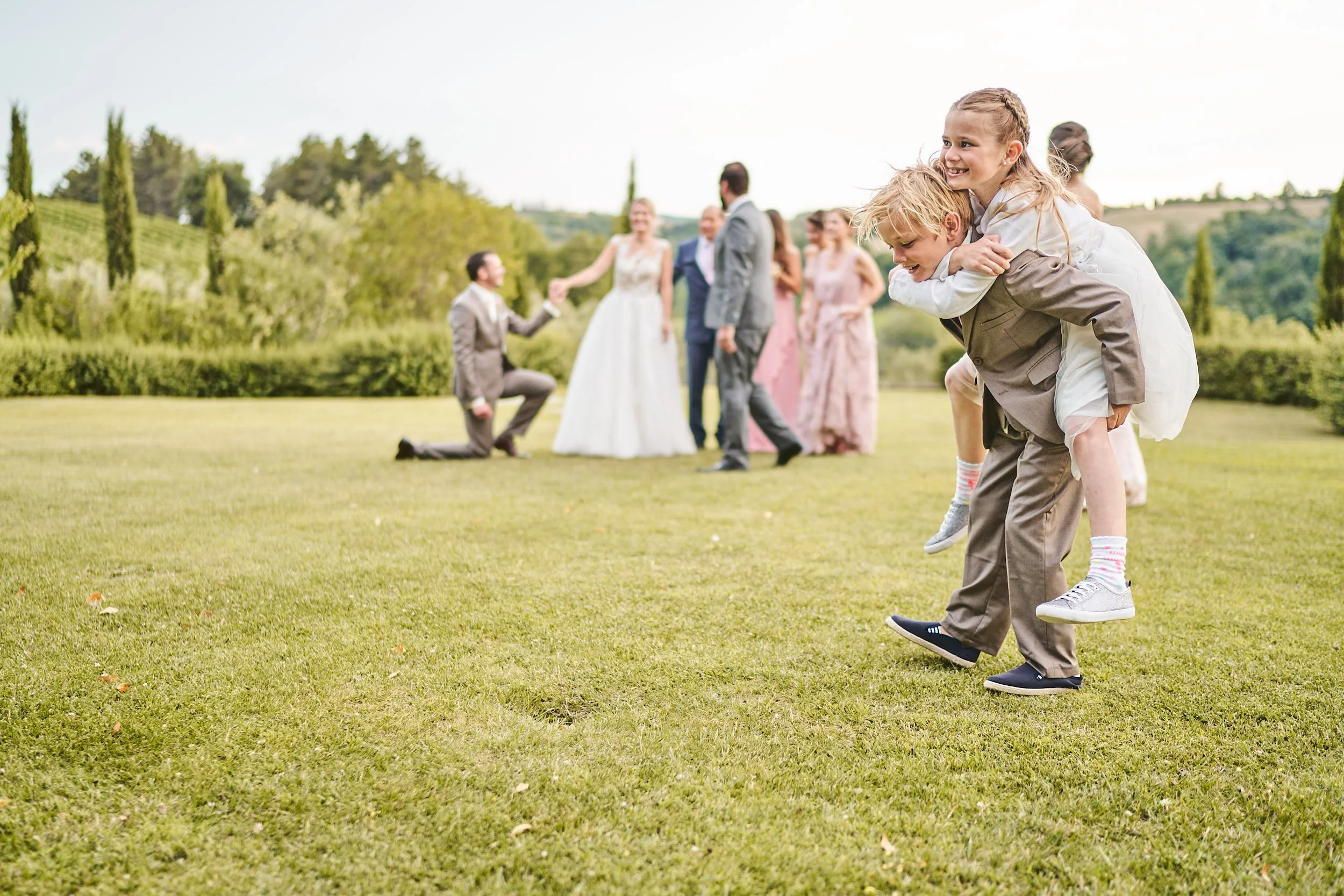Candid outdoor moment: a boy carries a girl piggyback across a lawn while the wedding party gathers in the background, playful and light.
