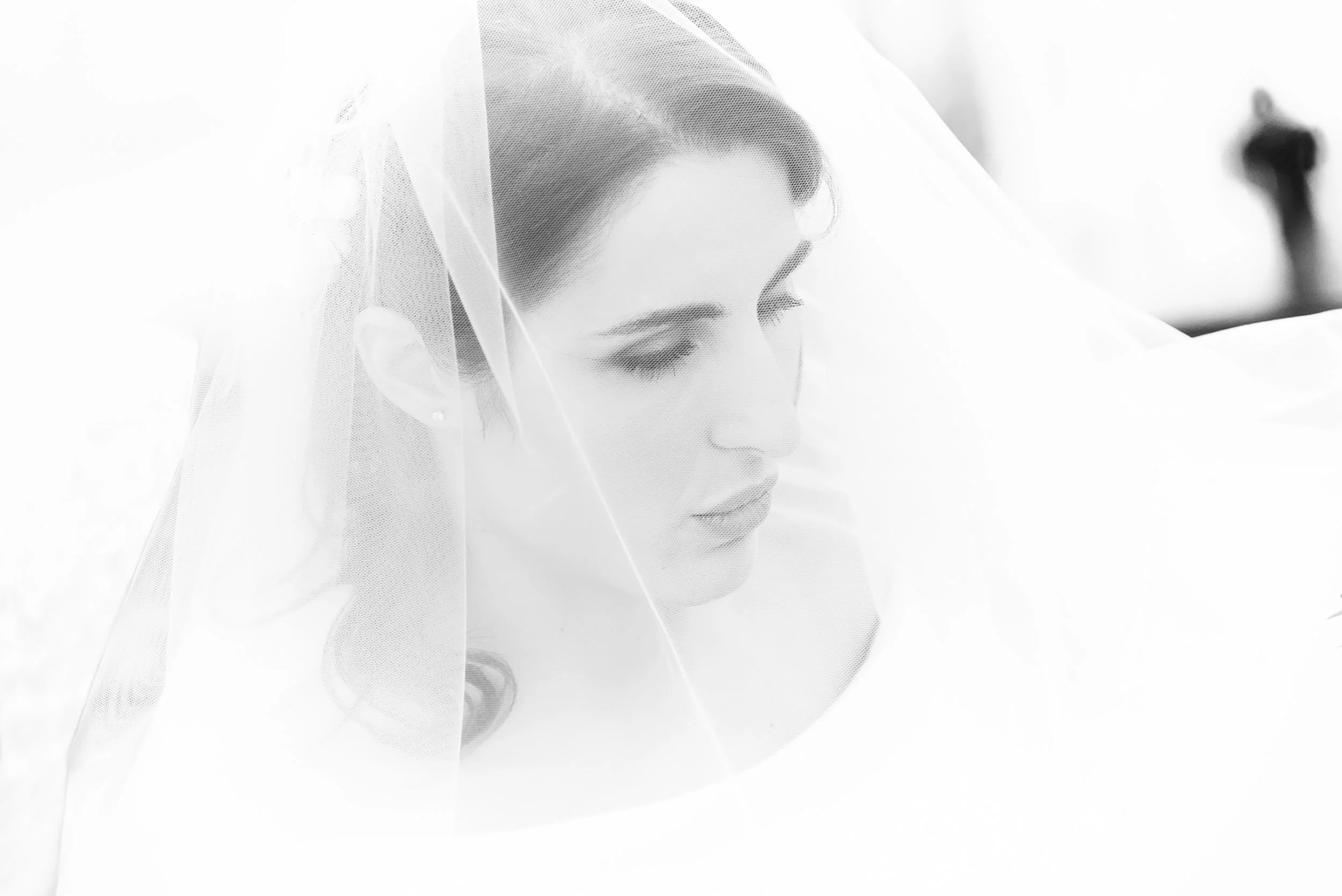 Minimal high-key bridal portrait framed by the veil, soft expression, bright airy background, with a blurred silhouette in the distance.