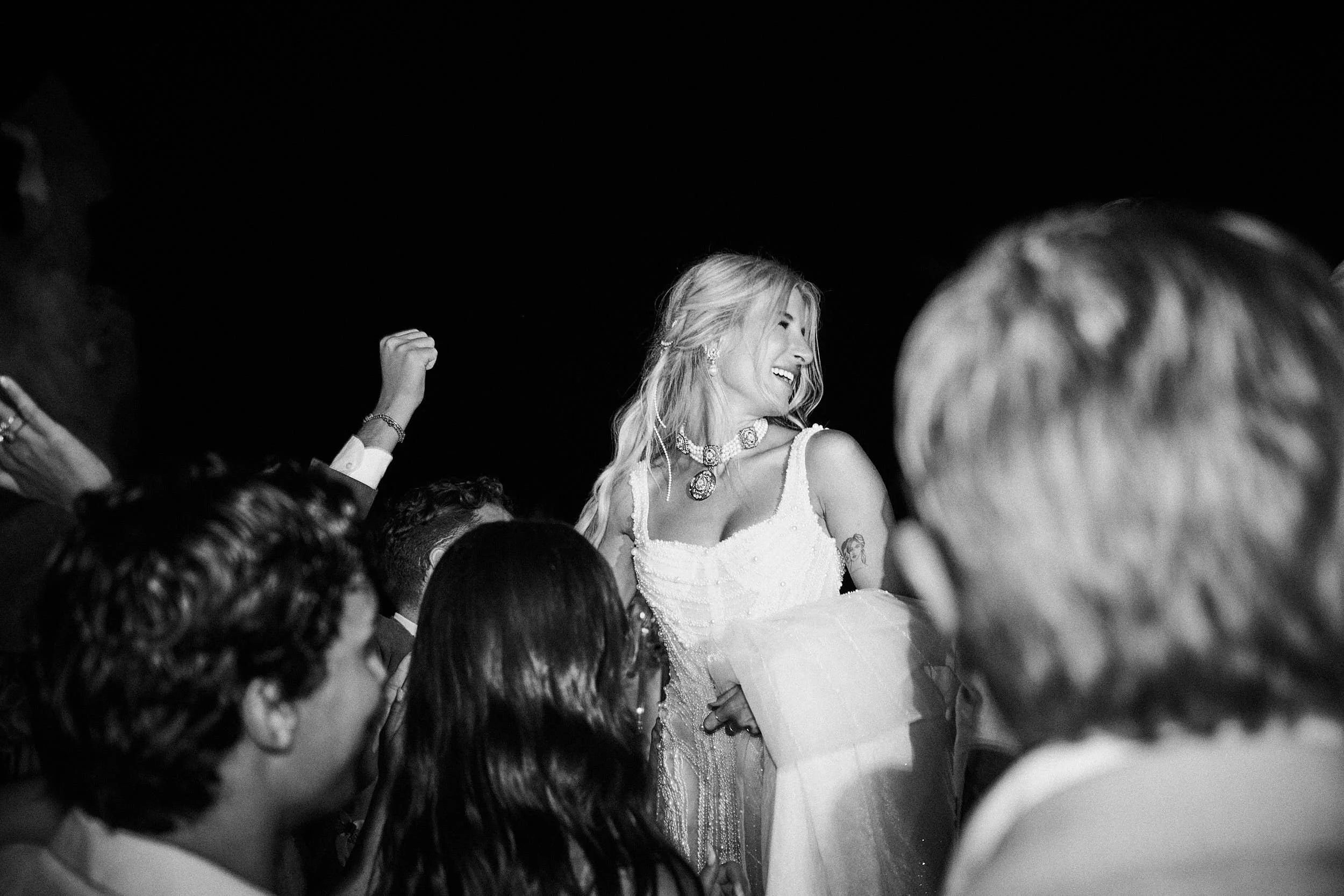 Black-and-white dance floor moment with the bride lifted in the crowd, captured mid-laughter during the party at The Rock, Phi Beach.