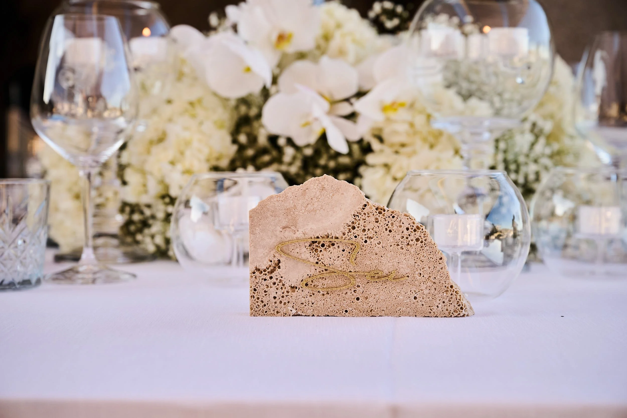 Stone place card detail on a white table setting, surrounded by glassware and white flowers, styled for a destination wedding reception in Sardinia.