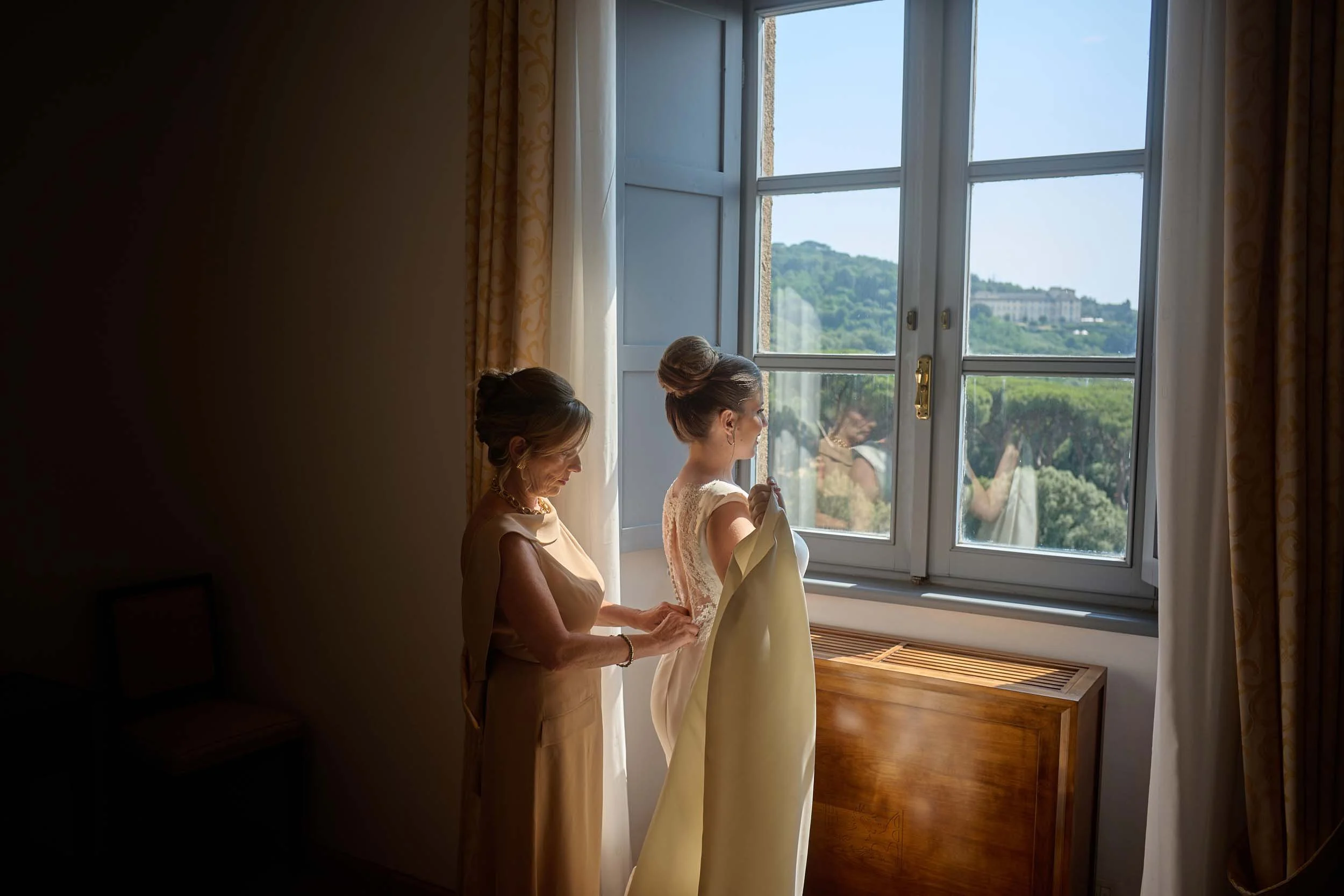 Bride’s mother buttons the back of her wedding dress at the window.