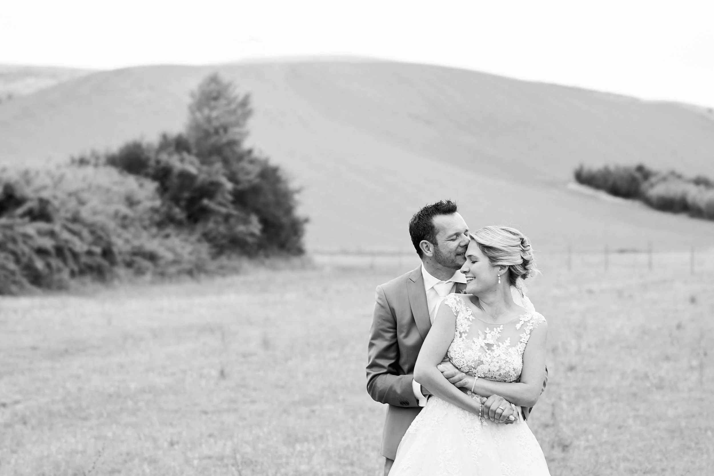 Bride and groom embracing in an open field, black and white portrait with soft hills in the background