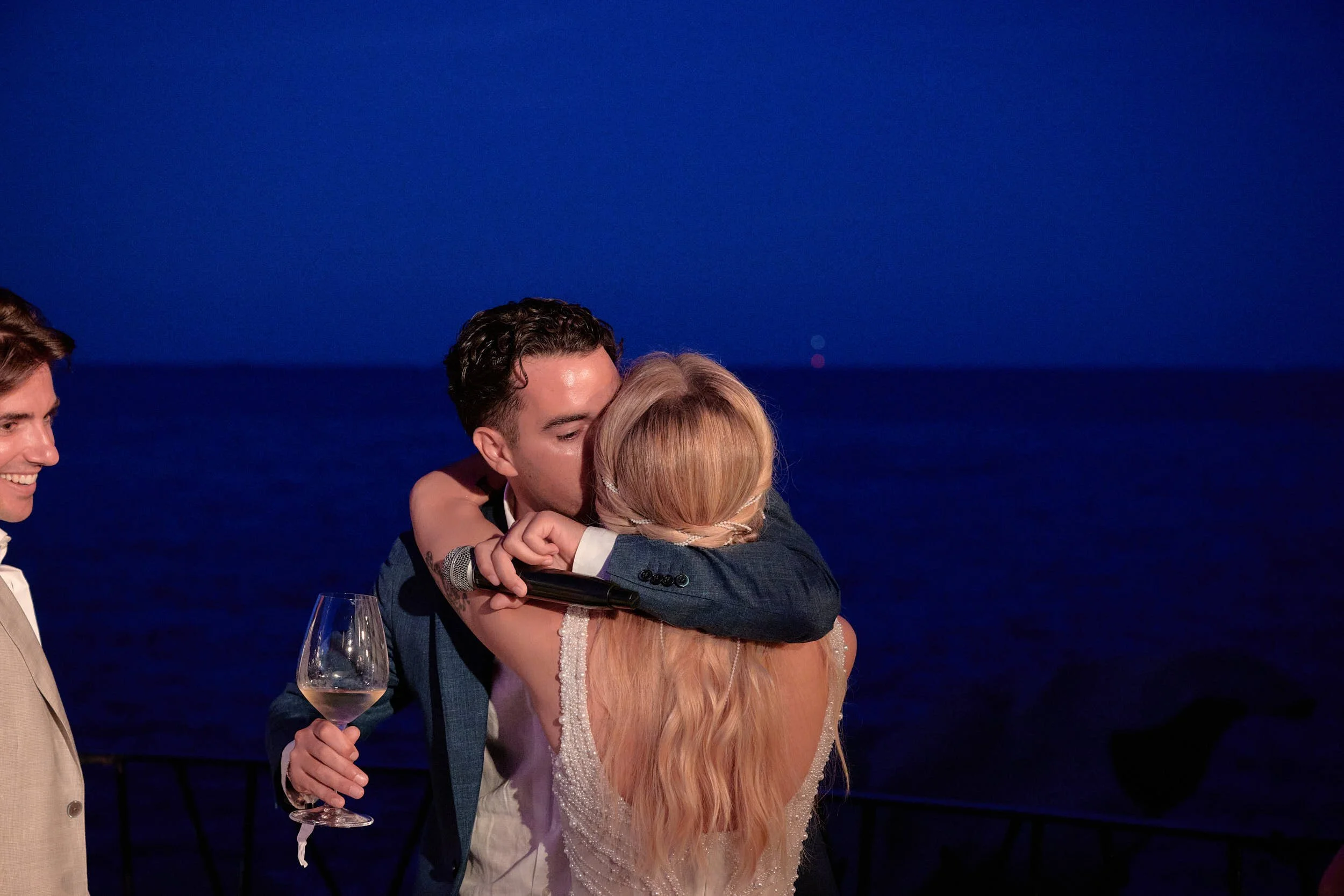 Bride and a friend embrace tightly during speeches, framed against the deep-blue sea at blue hour in Sardinia.
