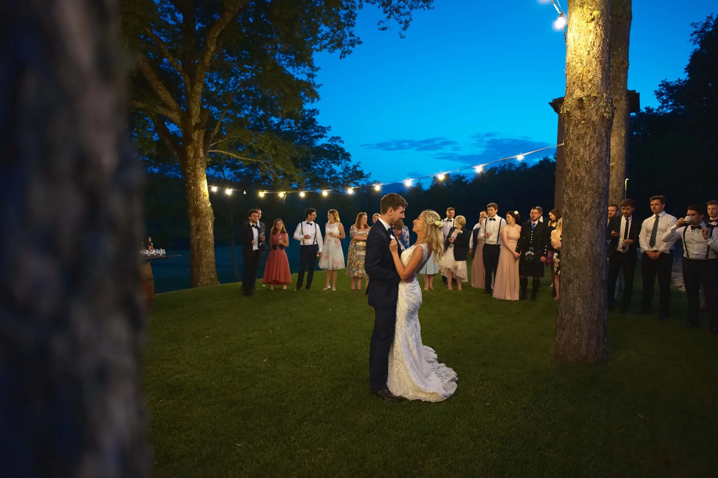 Bride and groom first dance under string lights in a Tuscan forest during blue hour — intimate wedding celebration