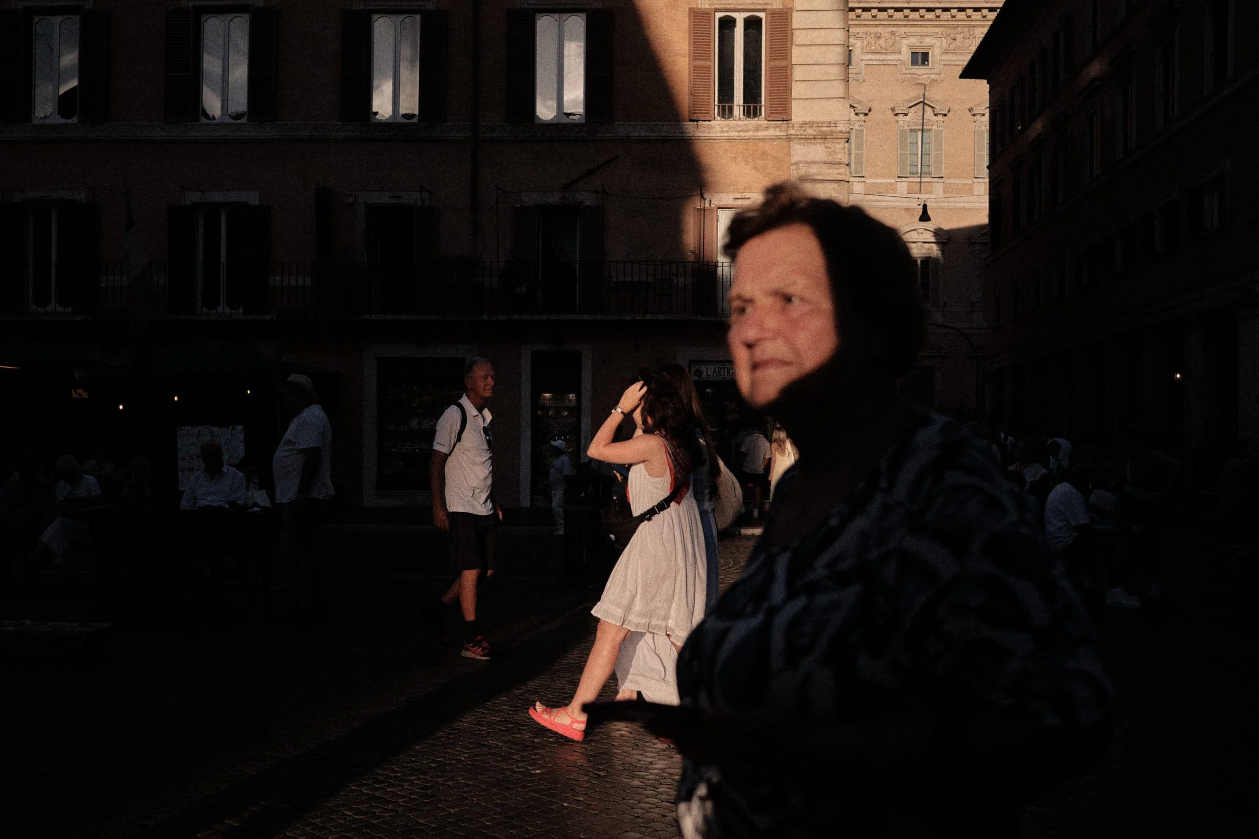 Street scene in Rome at golden hour, with an older woman in the foreground and pedestrians crossing a sunlit square in deep shadow.
