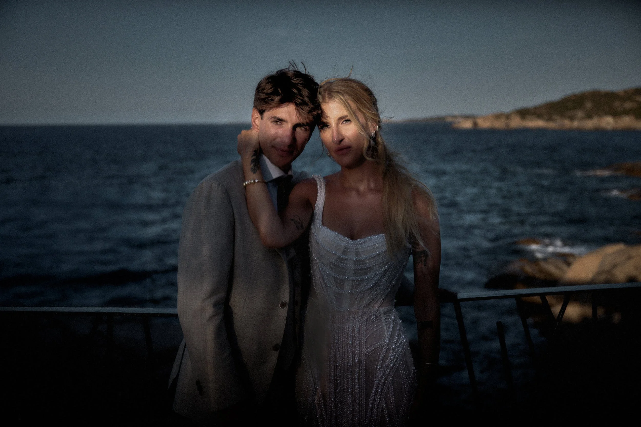 Intimate portrait of the newlyweds by the sea at blue hour, soft light on their faces and the coastline behind — destination wedding in Sardinia.