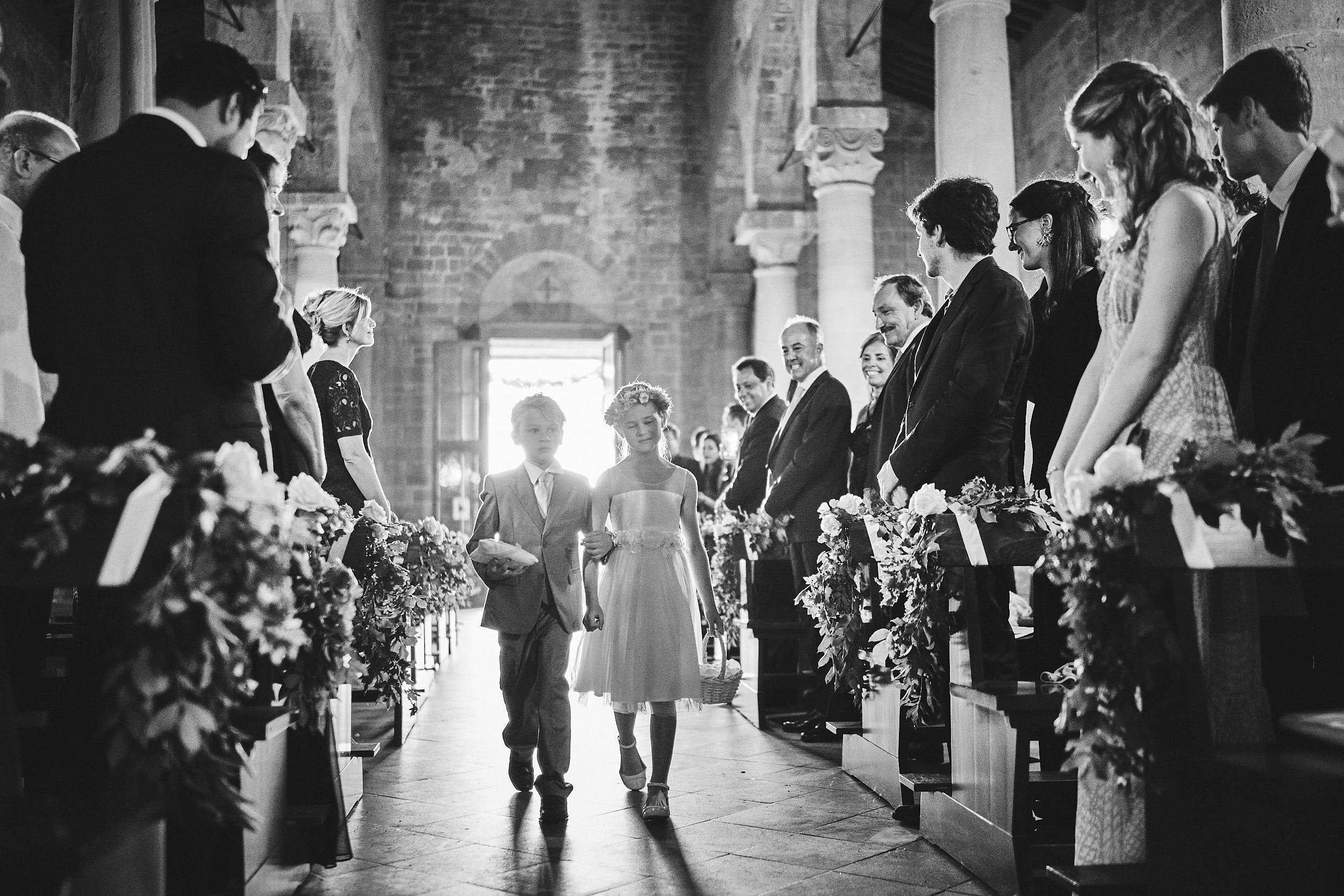 Two children walking down the aisle in a stone church, framed by guests and floral arrangements in soft light